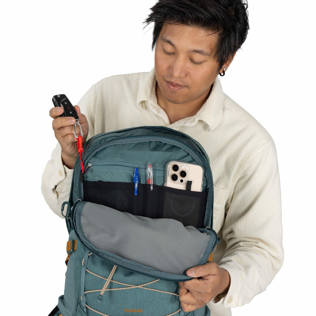 The image shows a person wearing a cream-colored long-sleeve shirt holding a teal-colored backpack from the brand "Quasar." The backpack is open, revealing its contents, which include a smartphone in one compartment and two pens in another, each slotting