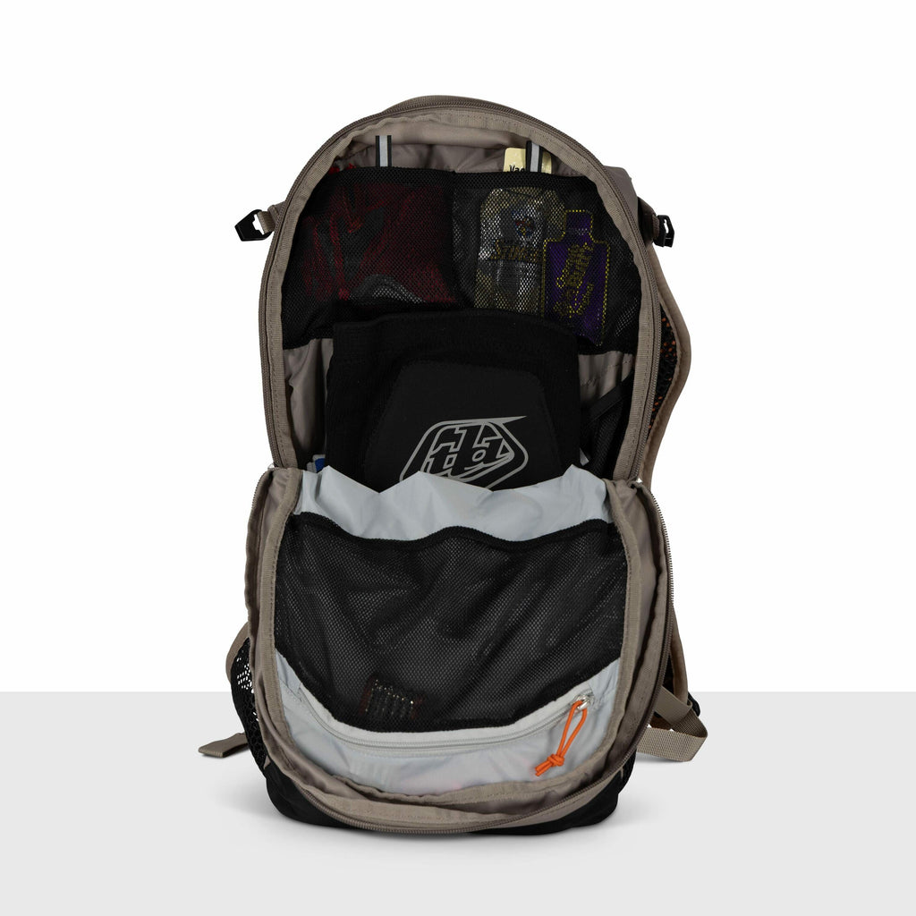 The image shows a backpack with its main compartment unzipped and open, revealing its contents. Inside, there's a black protective gear piece with a "TLD" logo, possibly a chest or back protector. Above it, there's a zippered mesh pocket containing variou