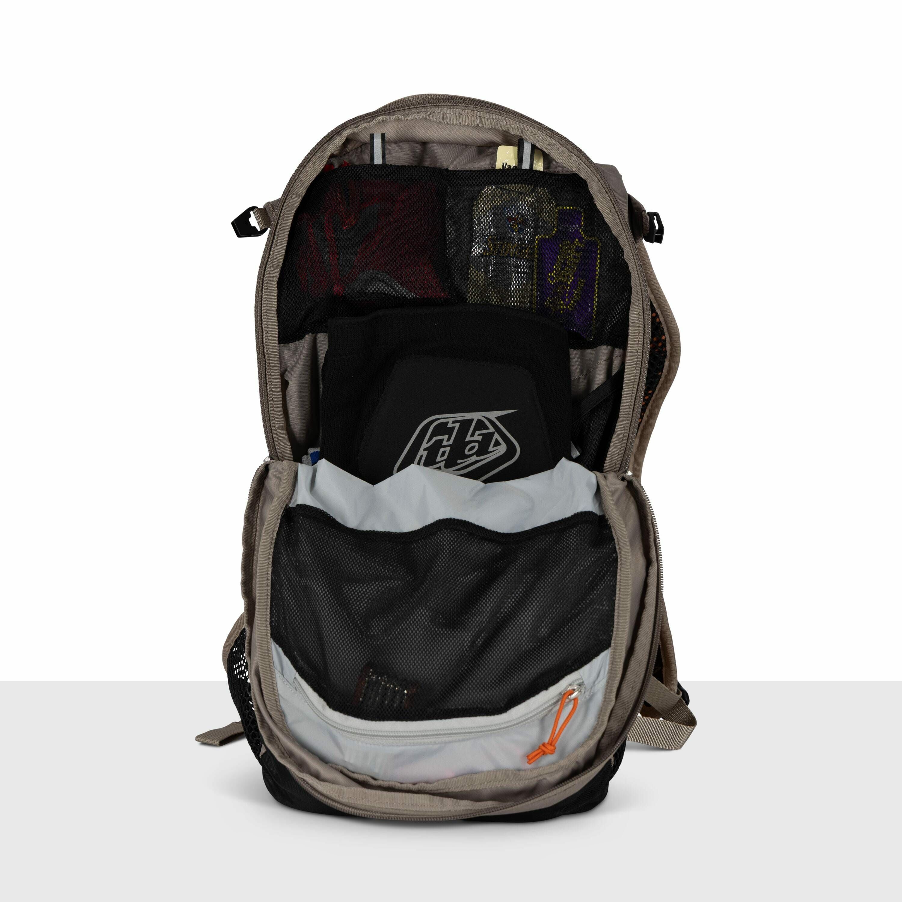 The image shows a backpack with its main compartment unzipped and open, revealing its contents. Inside, there's a black protective gear piece with a "TLD" logo, possibly a chest or back protector. Above it, there's a zippered mesh pocket containing variou