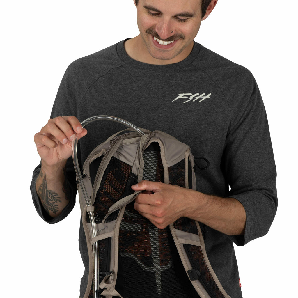 The image shows a person wearing a dark grey long-sleeve shirt with a "F14" logo on the chest. The person is holding a hydration backpack with their right hand and manipulating the drinking tube with their left hand. The backpack is grey and black, and th