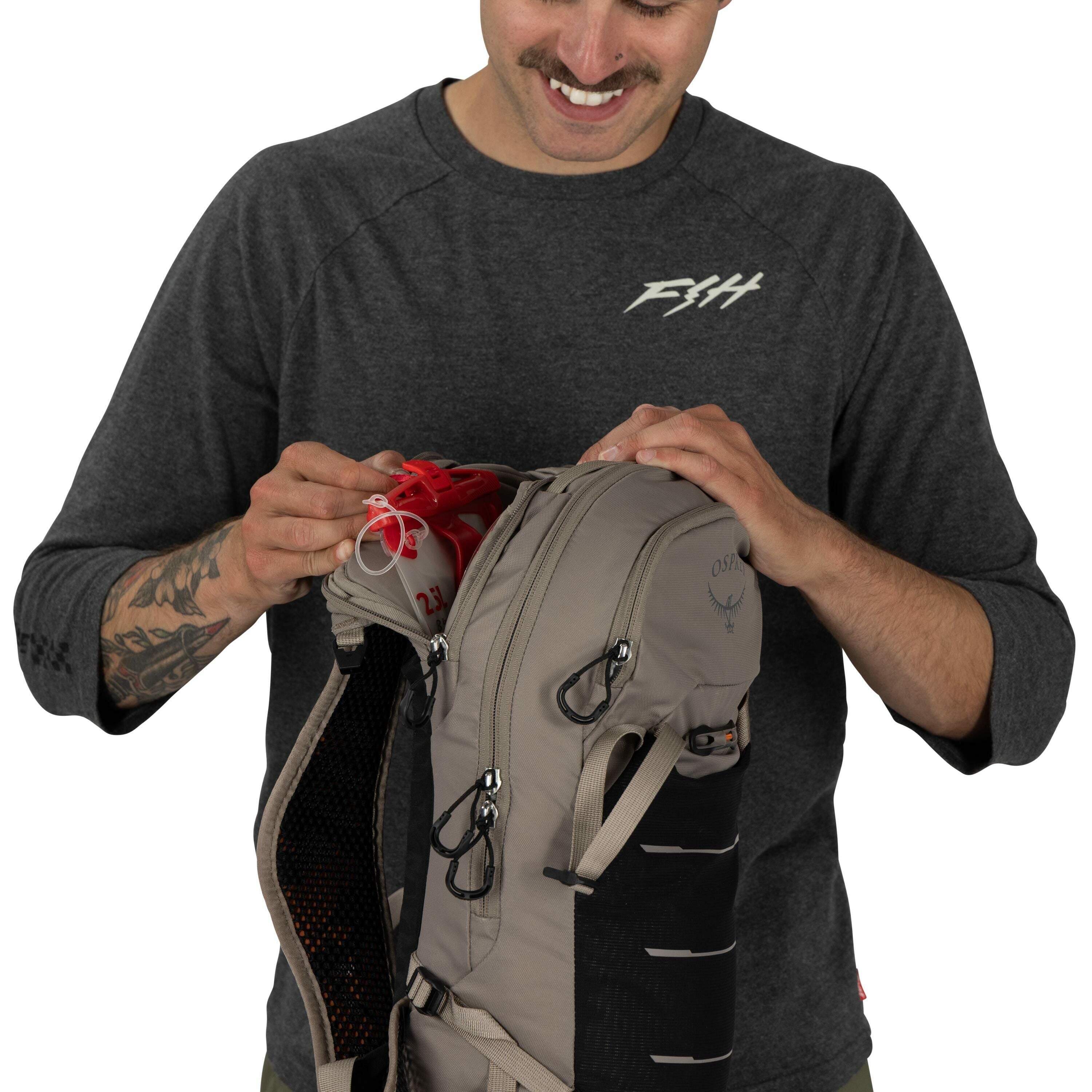 The image shows a person wearing a dark gray long-sleeve shirt with a logo "F&H" on the chest and a tattoo on their left forearm. They are handling a light gray backpack, which features black mesh side panels and multiple zippers and loops for attachments