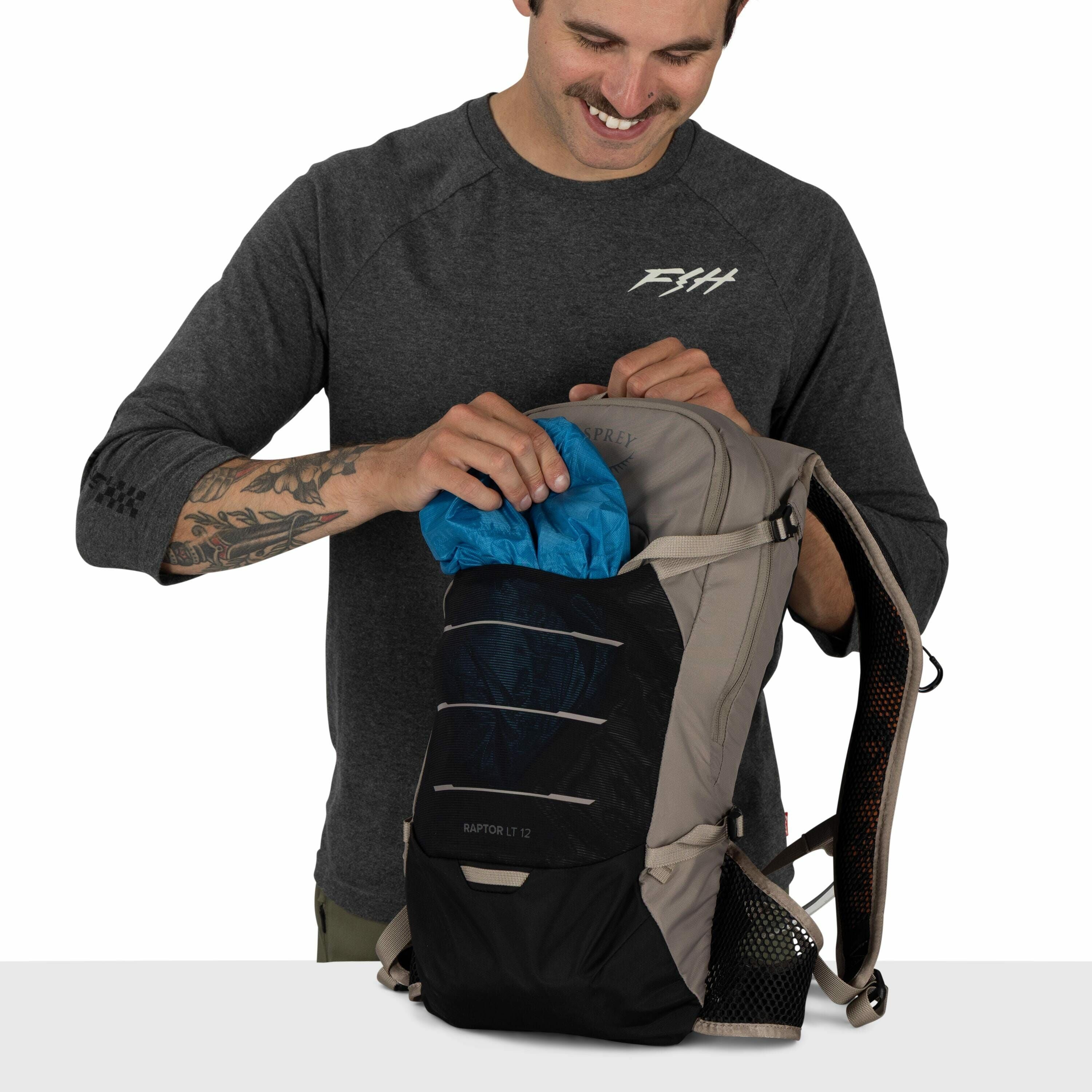 The image shows a person wearing a dark gray long-sleeve shirt with the logo "FSH" on the chest. The individual is holding a light gray backpack with black accents and the logo "Osprey" visible at the top. The backpack model, "Raptor LT 12," is written on
