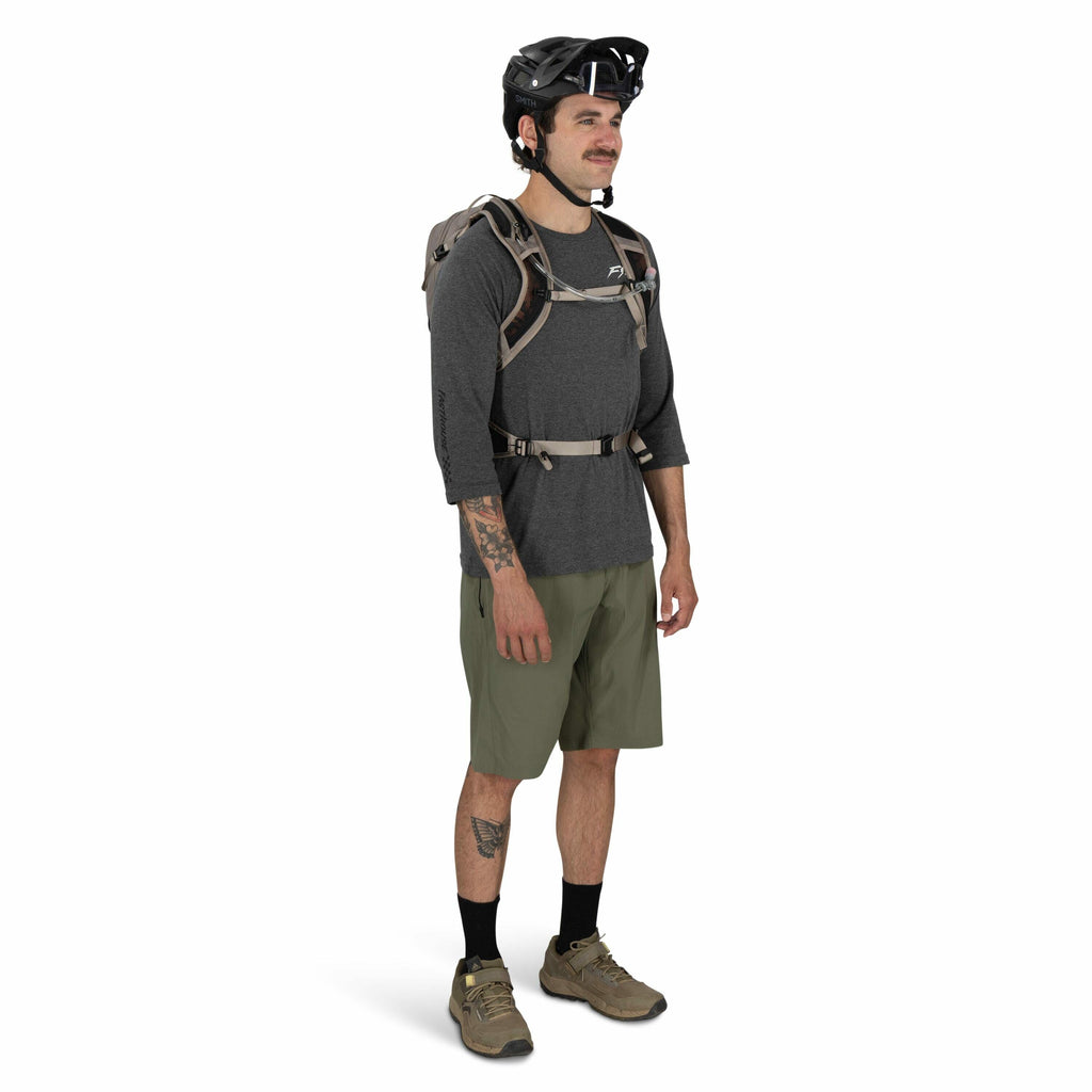 The image features an individual dressed in casual outdoor attire. The person is wearing a gray long-sleeved shirt with a logo on the chest area and olive green shorts. They have a tattoo on their left forearm and a tattoo on their left calf. The individu