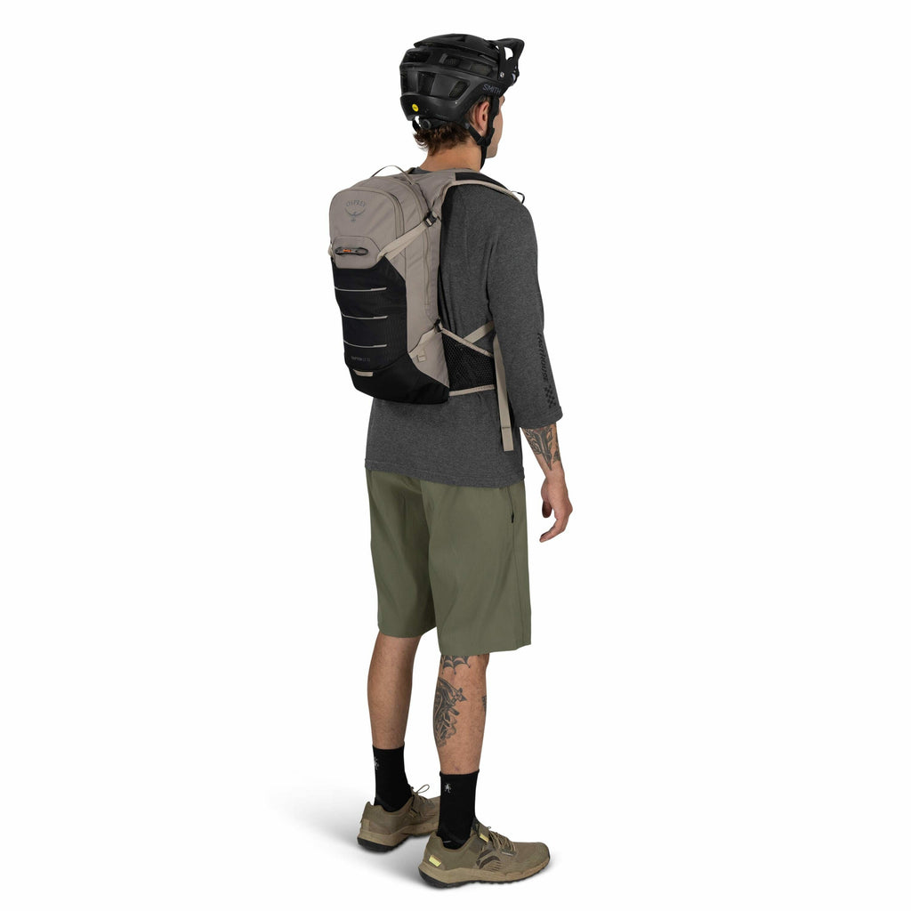 The image shows a man dressed for outdoor activity, likely mountain biking or hiking, wearing a helmet and a backpack. The man is standing with his back slightly turned towards the camera, showcasing the backpack on his back. He is wearing a grey long-sle