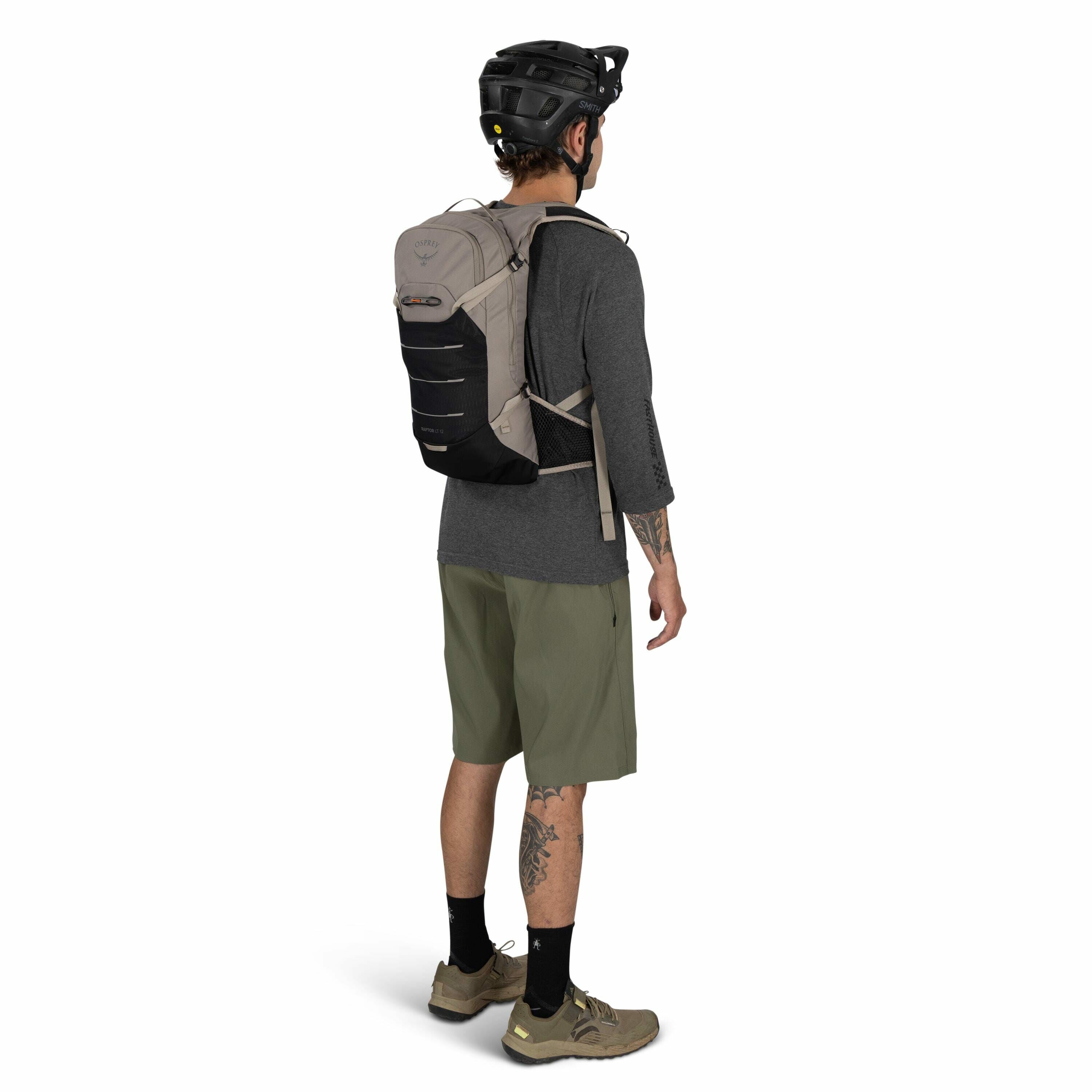 The image shows a man dressed for outdoor activity, likely mountain biking or hiking, wearing a helmet and a backpack. The man is standing with his back slightly turned towards the camera, showcasing the backpack on his back. He is wearing a grey long-sle
