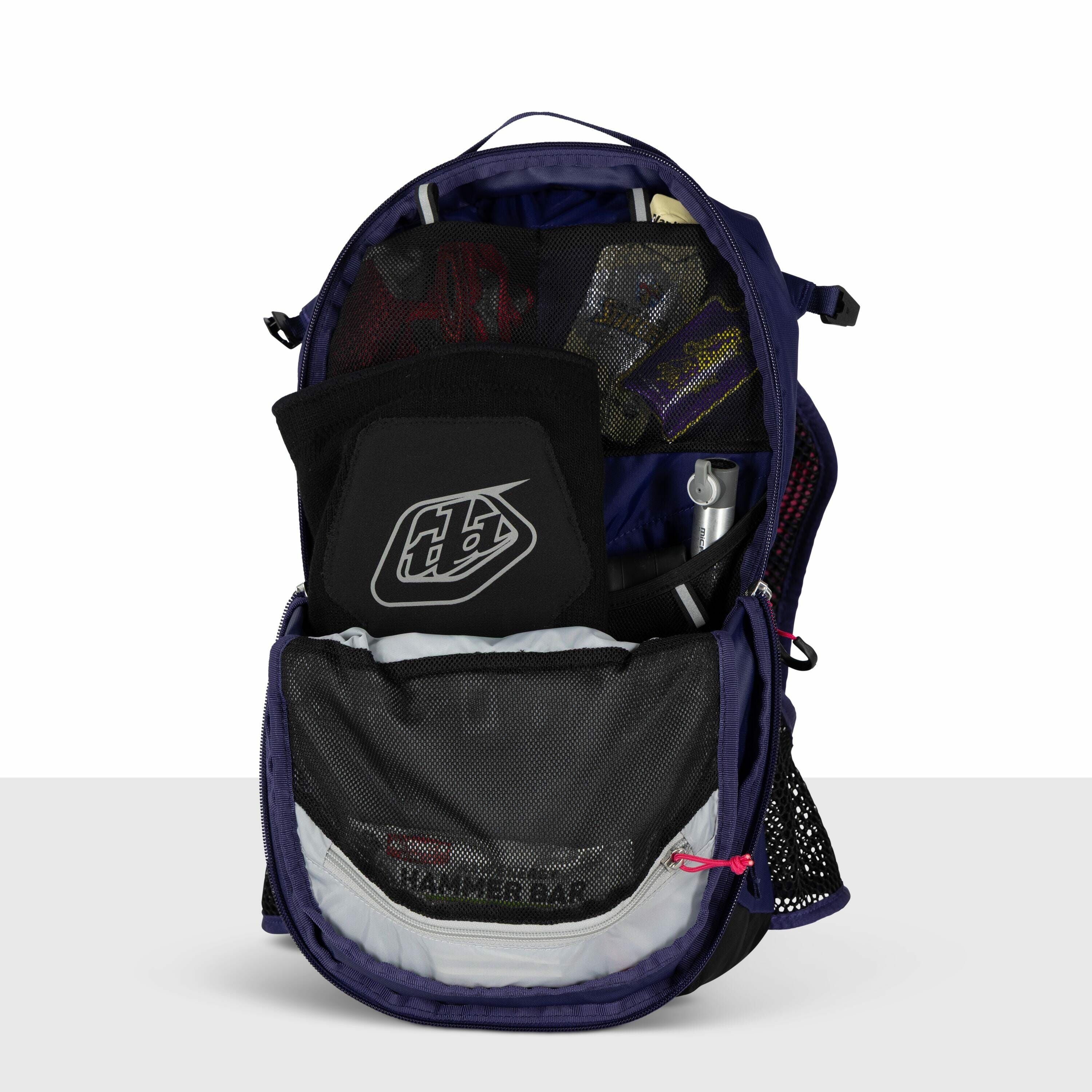 The image shows an open backpack, displaying its contents. Inside the backpack, there is a black protective pad with a white logo that reads "Troy Lee Designs." There are also several small items tucked into mesh pockets, including what appears to be pack