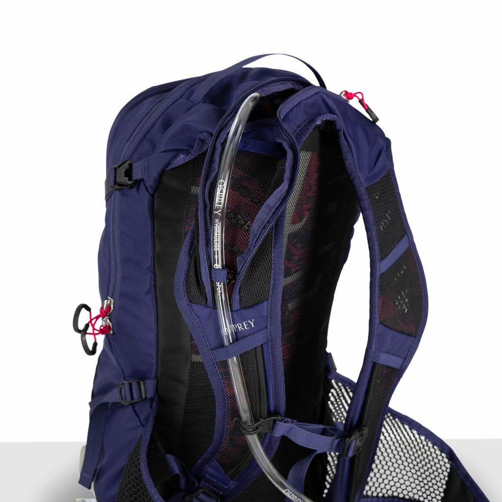 The image features a close-up view of a blue Osprey backpack, focusing on its hydration system. A transparent hydration tube is neatly threaded through a loop on the backpack's shoulder strap, making it easily accessible to the user. The backpack has padd