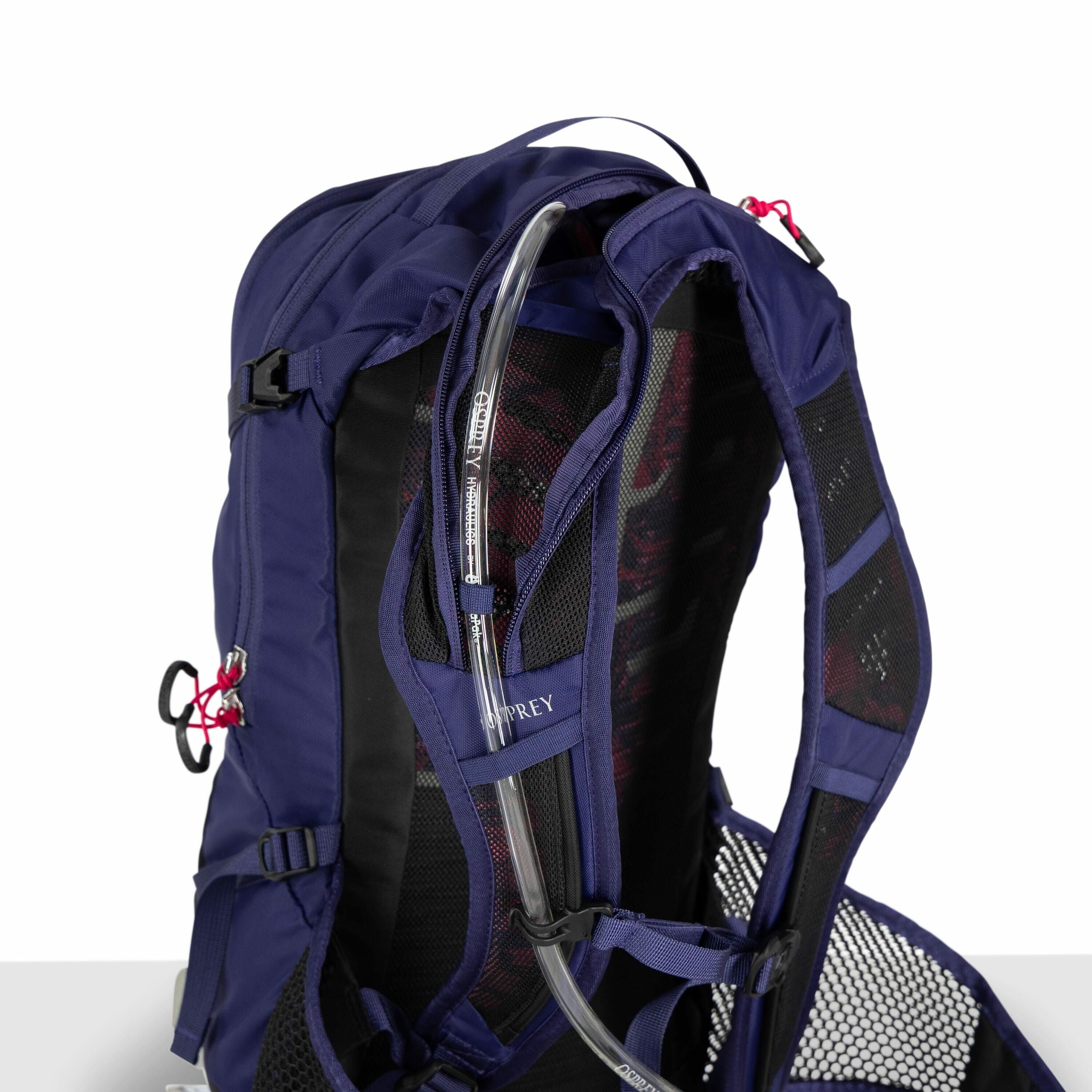 The image features a close-up view of a blue Osprey backpack, focusing on its hydration system. A transparent hydration tube is neatly threaded through a loop on the backpack's shoulder strap, making it easily accessible to the user. The backpack has padd
