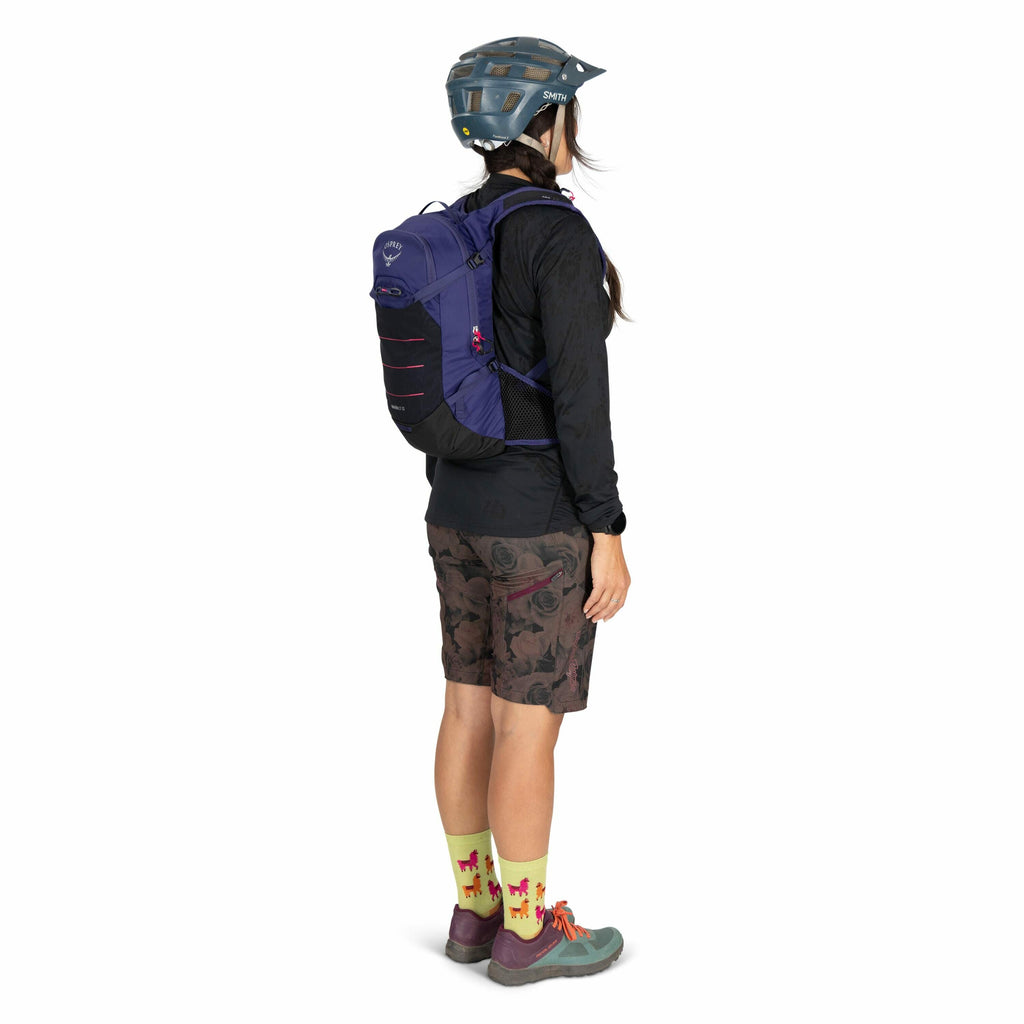 The image depicts a woman standing in profile, wearing a blue Smith helmet and a blue Osprey backpack. She is dressed in a long-sleeve black top and knee-length shorts with a dark floral pattern. Her socks are bright yellow with a playful design featuring