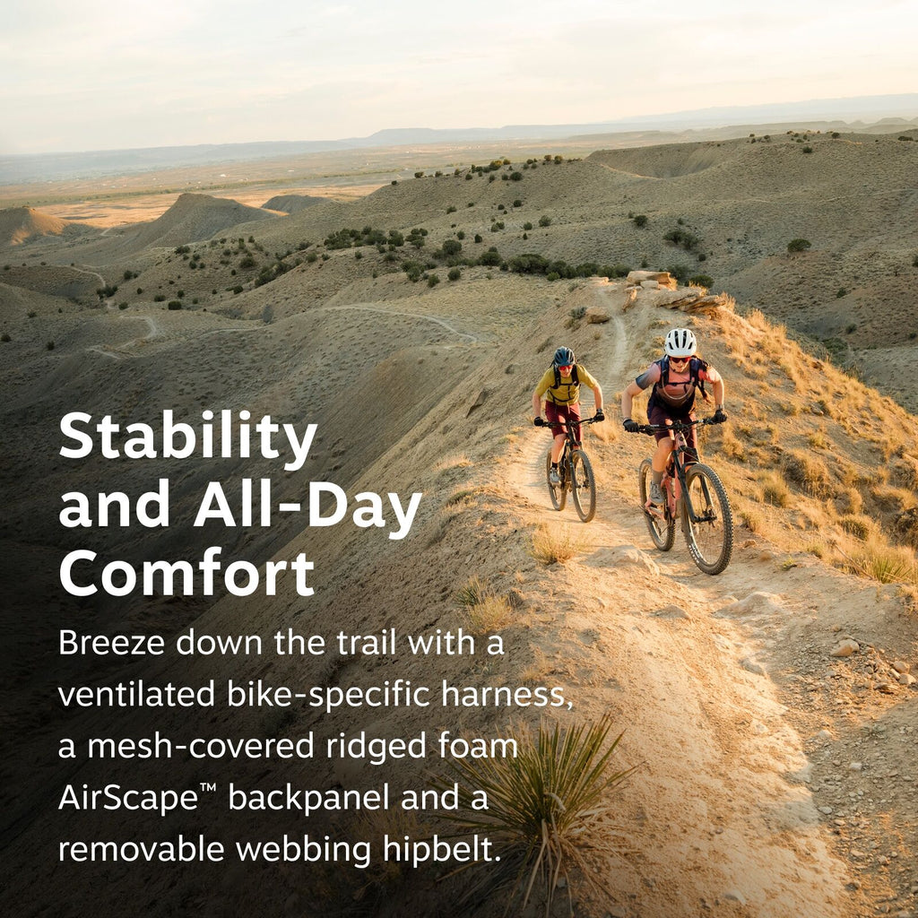 The image captures two mountain bikers riding along a narrow, dusty trail on a ridge with expansive desert-like terrain in the background. The scene is set during golden hour, evident from the warm tones of the sunlight casting long shadows across the tra
