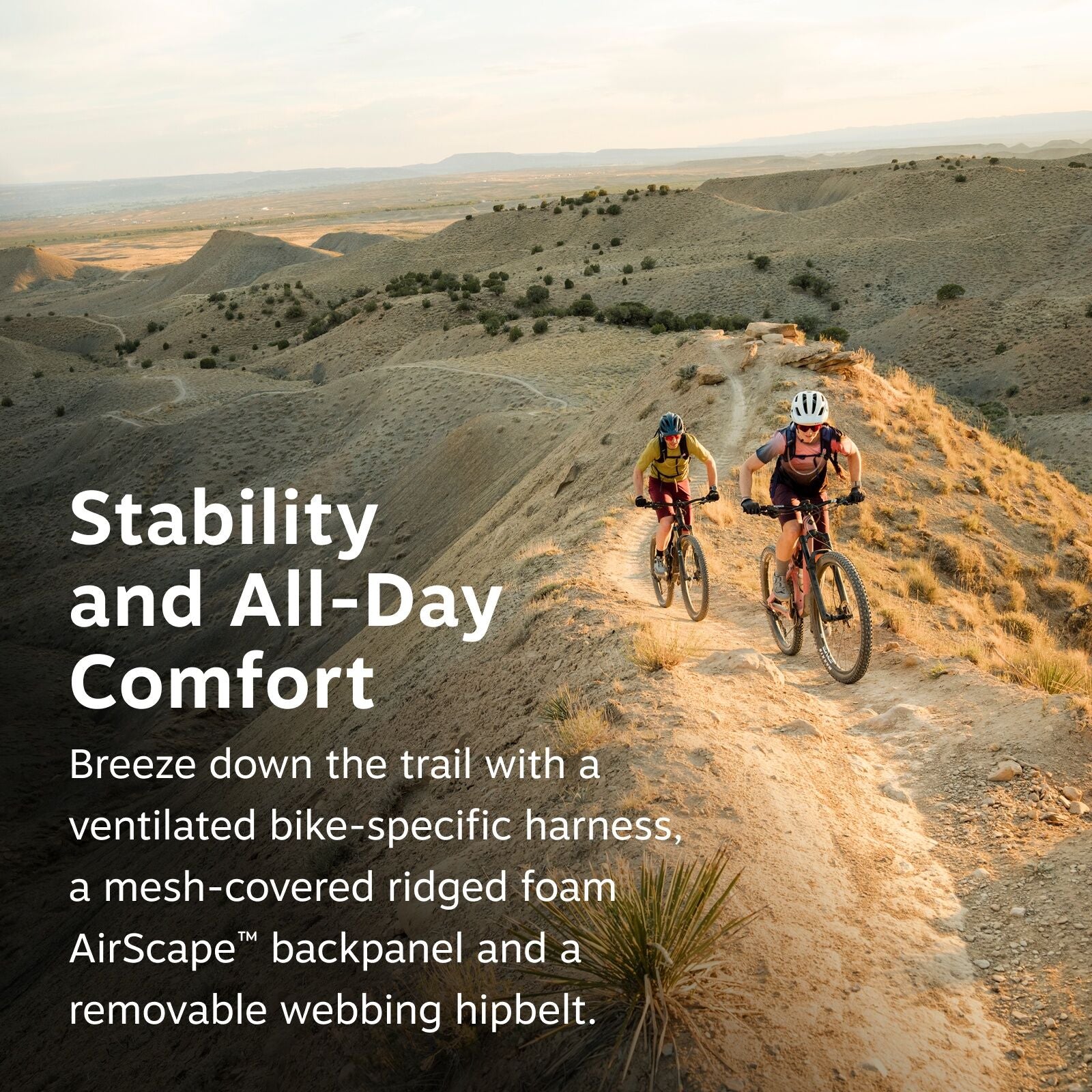 The image captures two mountain bikers riding along a narrow, dusty trail on a ridge with expansive desert-like terrain in the background. The scene is set during golden hour, evident from the warm tones of the sunlight casting long shadows across the tra