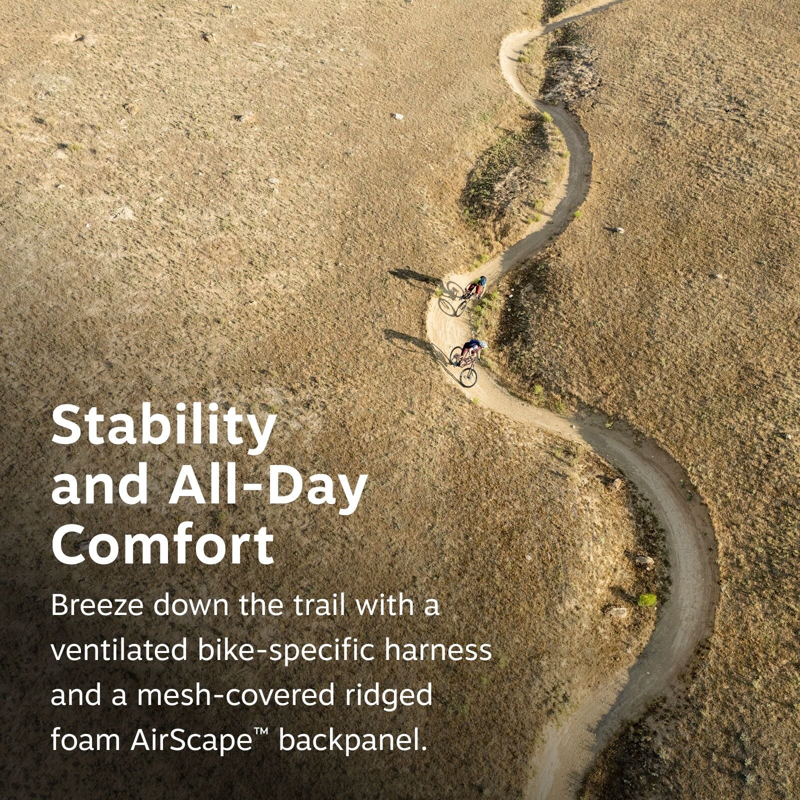 The image depicts two mountain bikers riding along a winding dirt trail on a grassy hillside. The trail snakes back and forth, creating a dynamic zigzag pattern as it traverses the natural landscape. The scene is captured from an aerial perspective, offer