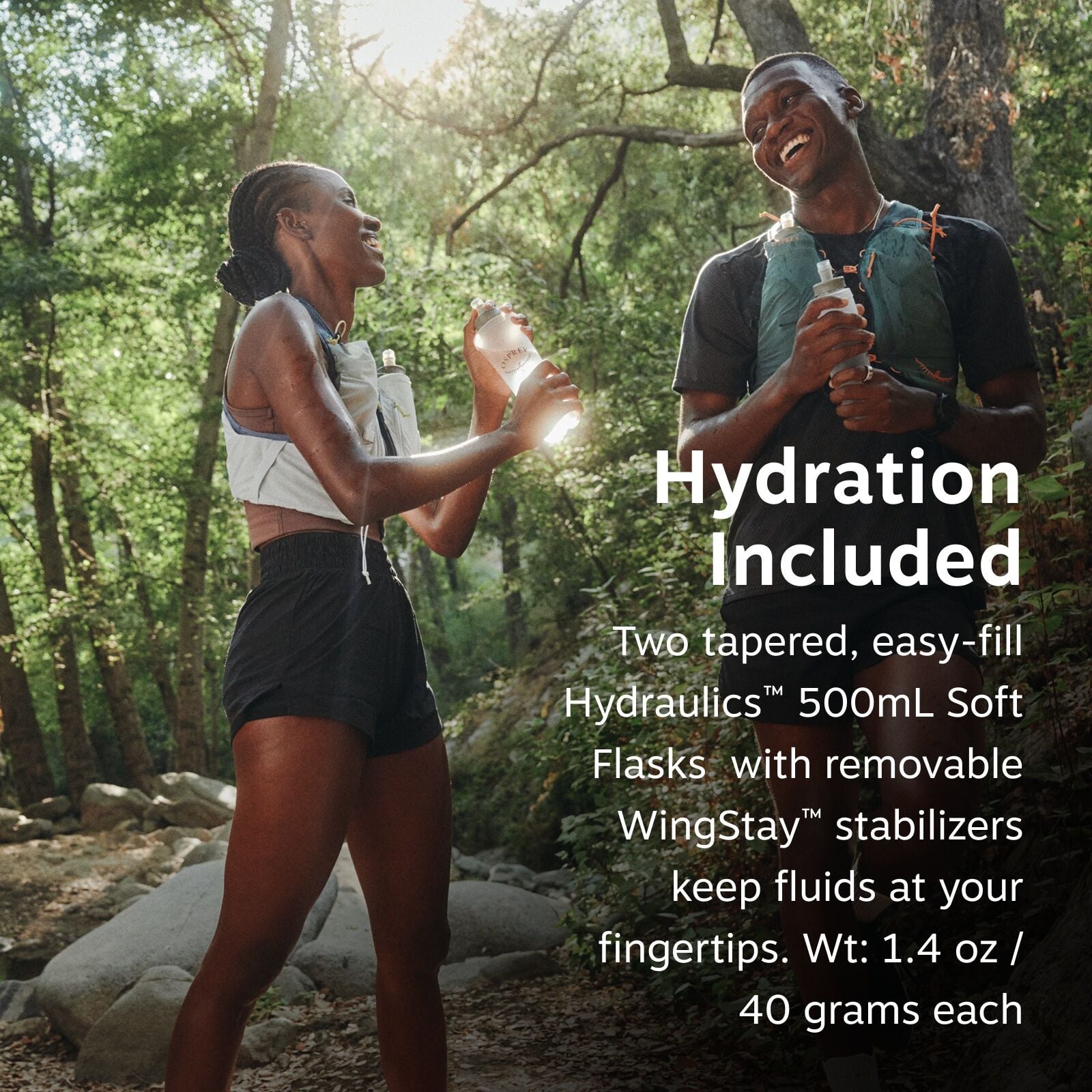 The image depicts two people in athletic attire standing in a forested area, each holding a hydration flask. They appear to be engaged in an outdoor activity, likely hiking or trail running, as indicated by their gear and the natural surroundings. Sunligh
