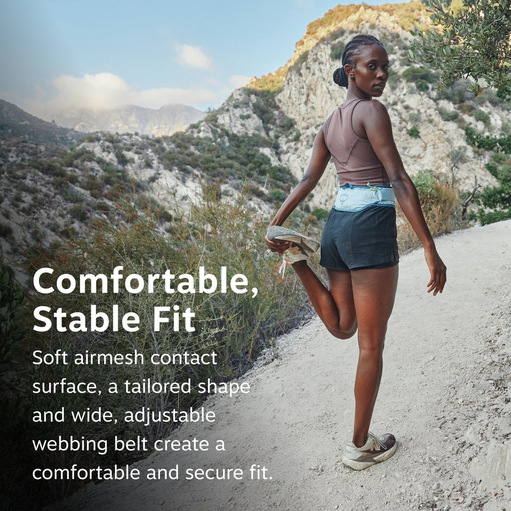 The image features a person standing on a hiking trail surrounded by mountainous terrain. The person is stretching one leg back while holding onto their foot, indicating a warm-up or cool-down exercise during a hike. They are wearing a brown sleeveless to