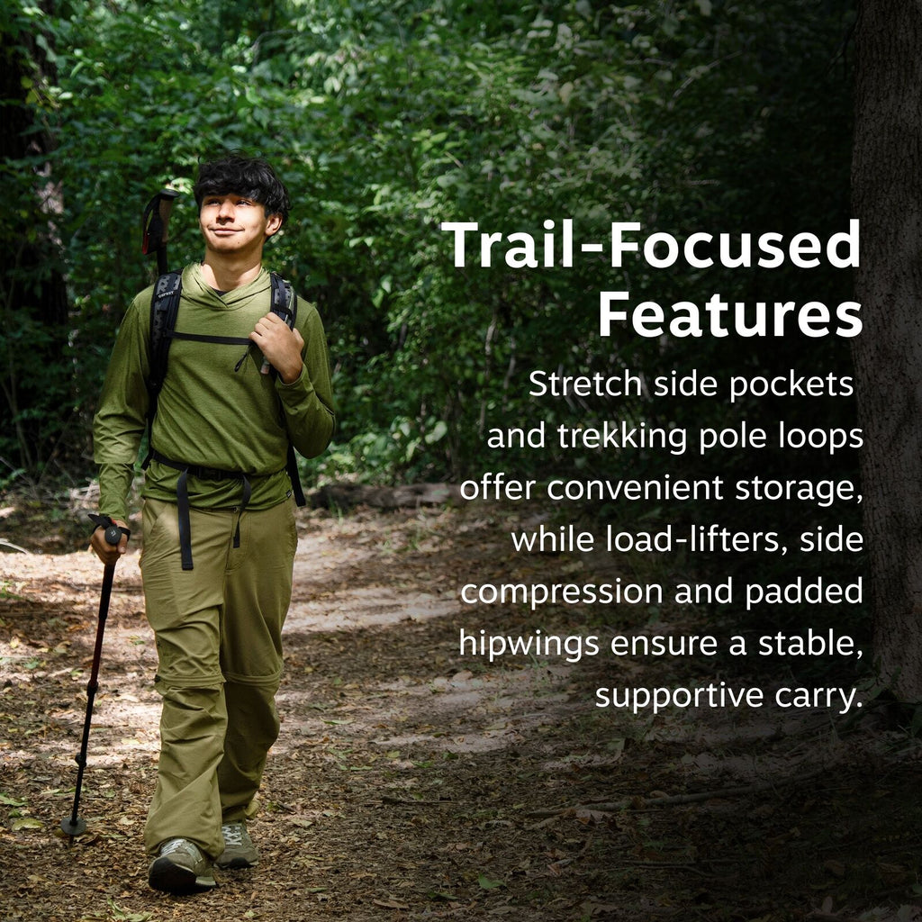 The image features an individual hiking on a forest trail, surrounded by lush green foliage. The person is equipped with a backpack and holding a trekking pole in one hand. They are dressed in outdoor gear, including a long-sleeved green shirt and khaki p