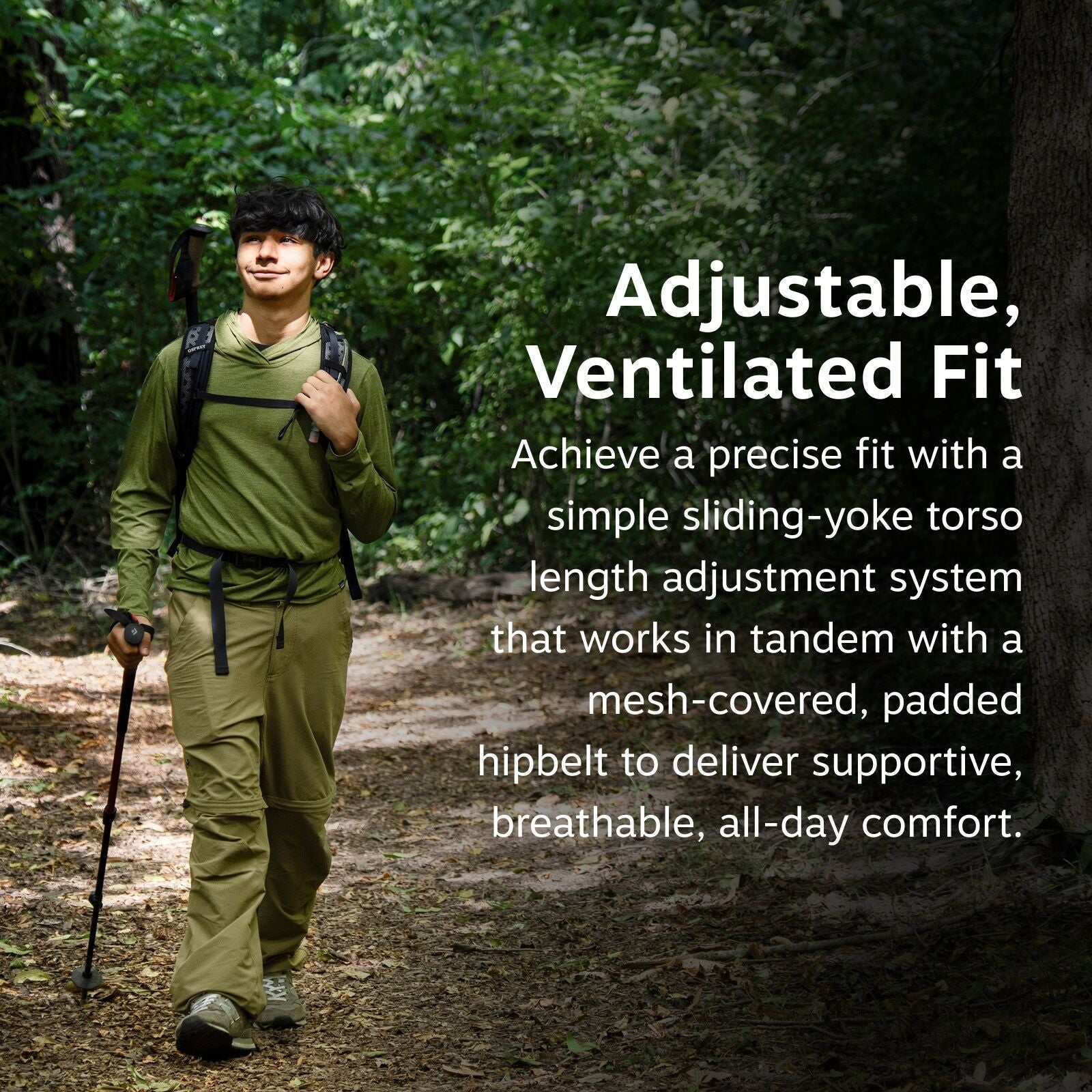 The image features a person walking through a wooded area, dressed in outdoor hiking attire. They are wearing a green long-sleeved shirt and khaki pants, equipped with a backpack. The person is using a hiking pole to aid their walk on the forest path. The