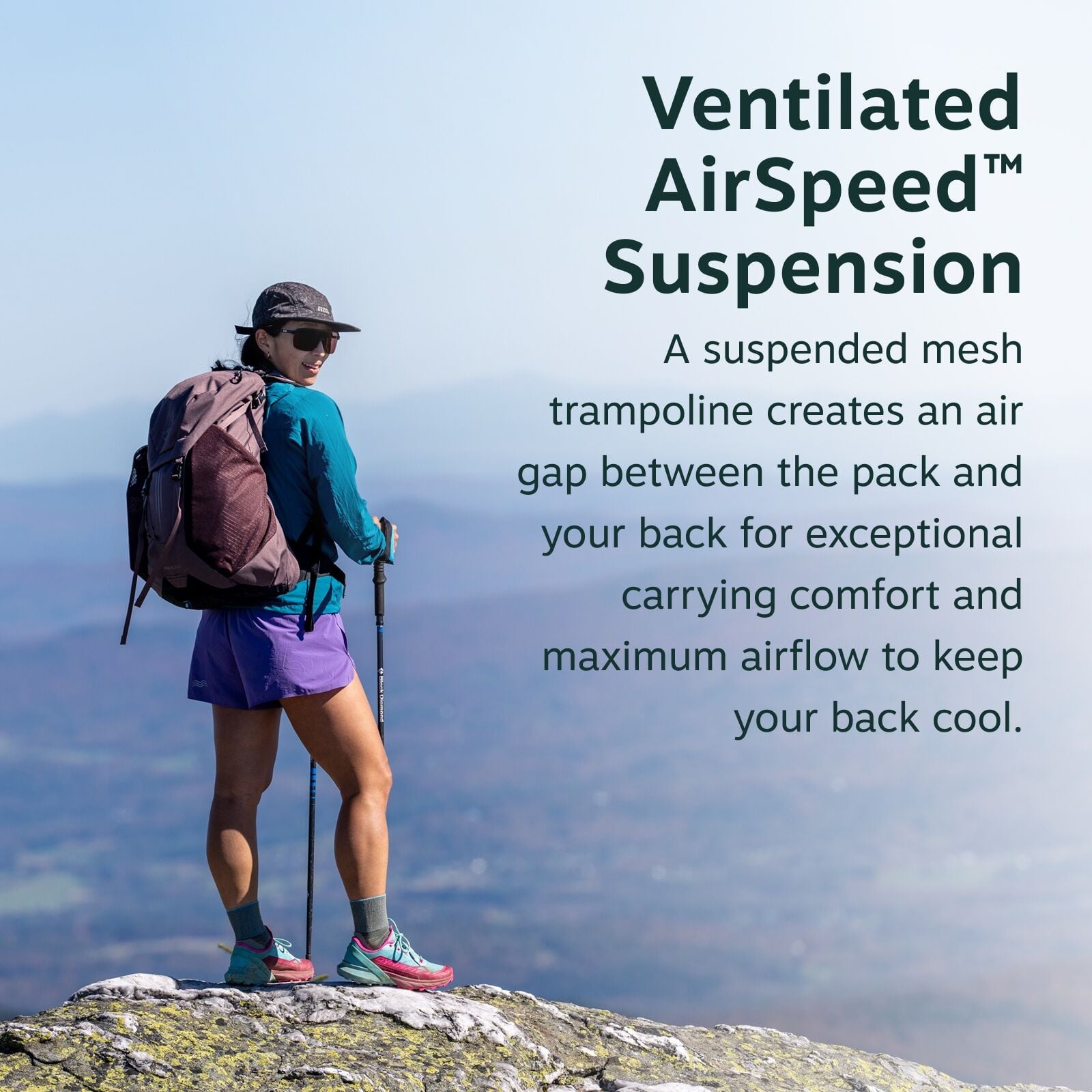The image shows a person standing on a rocky outcrop, equipped for hiking. They are wearing a blue long-sleeve shirt and purple shorts, complemented by hiking shoes, socks, and a cap. The individual carries a large brown backpack and holds hiking poles, g