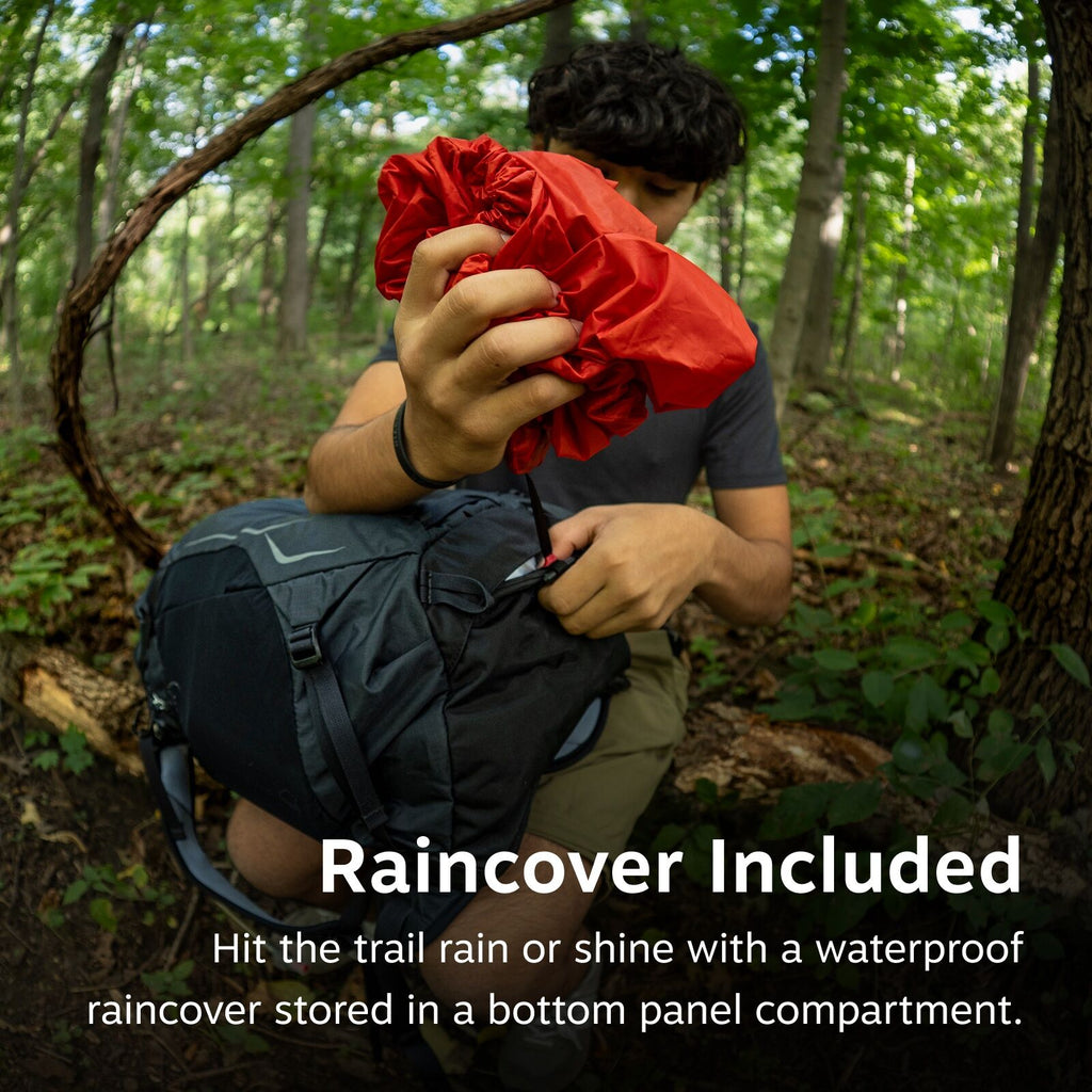 The image shows a person in a forest setting, holding a bright red rain cover. They appear to be taking it out from a bottom compartment of a dark-colored backpack. The scene is lush with green foliage, indicating a dense forest environment. The backpack