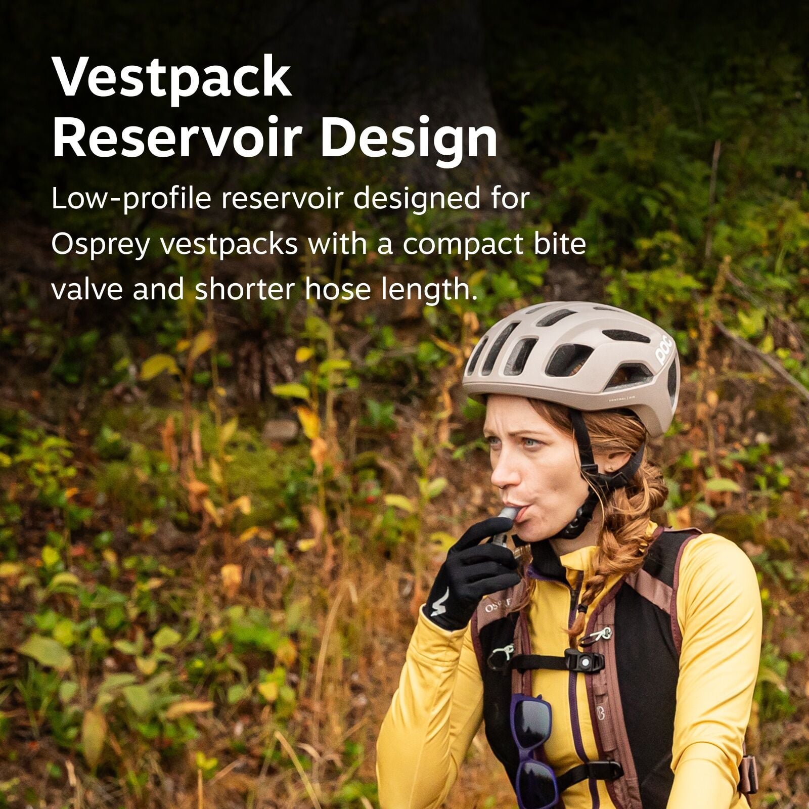 The image shows an individual in a natural, outdoor setting wearing cycling gear, including a protective POC helmet. They are also dressed in a yellow long-sleeve shirt with a black vestpack that features a hydration system. The vestpack appears to have a