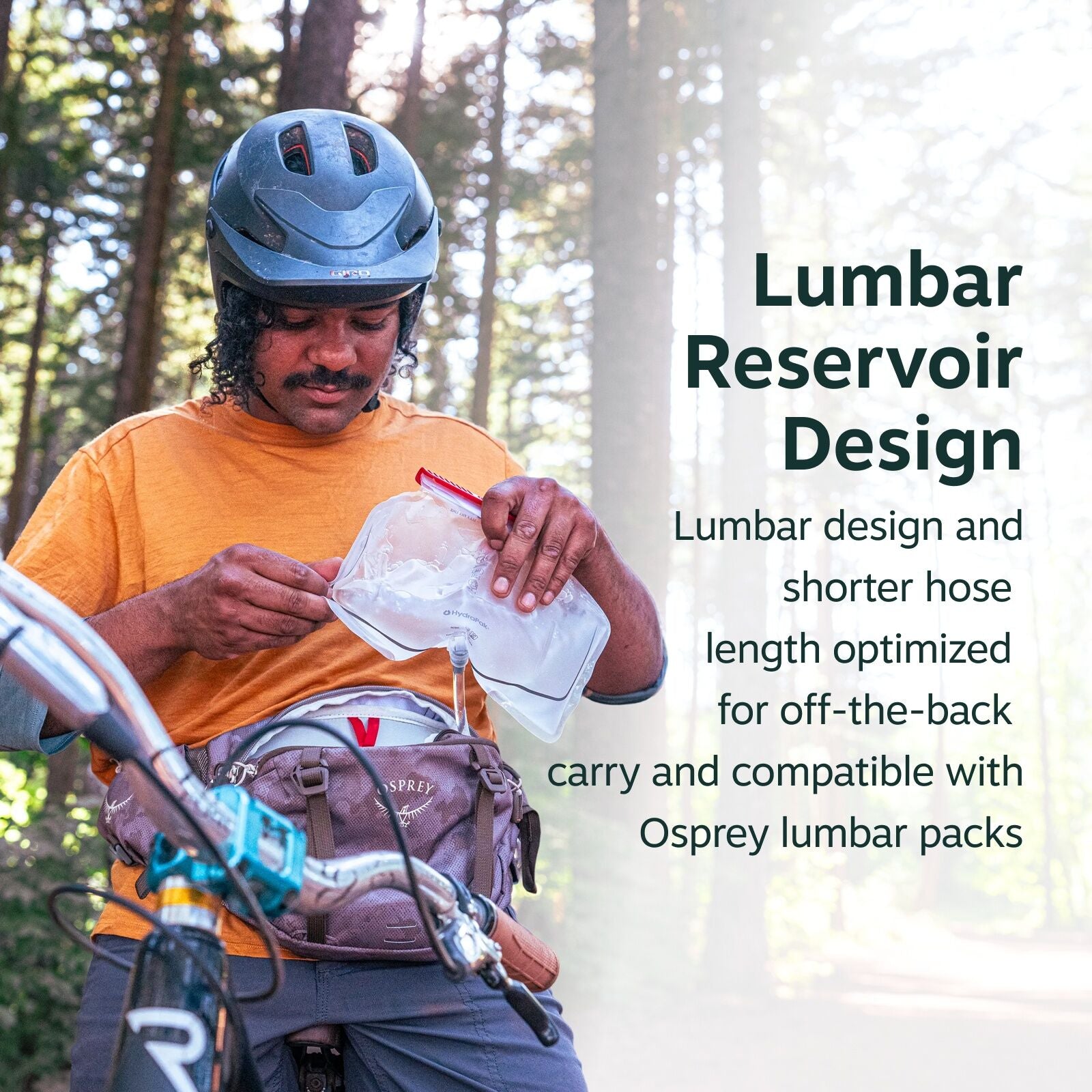 The image shows a person standing with a bicycle in a forested area, wearing a blue helmet and an orange T-shirt. They are holding a transparent hydration reservoir with a red cap, which is designed to fit in a lumbar pack. The Osprey lumbar pack around t