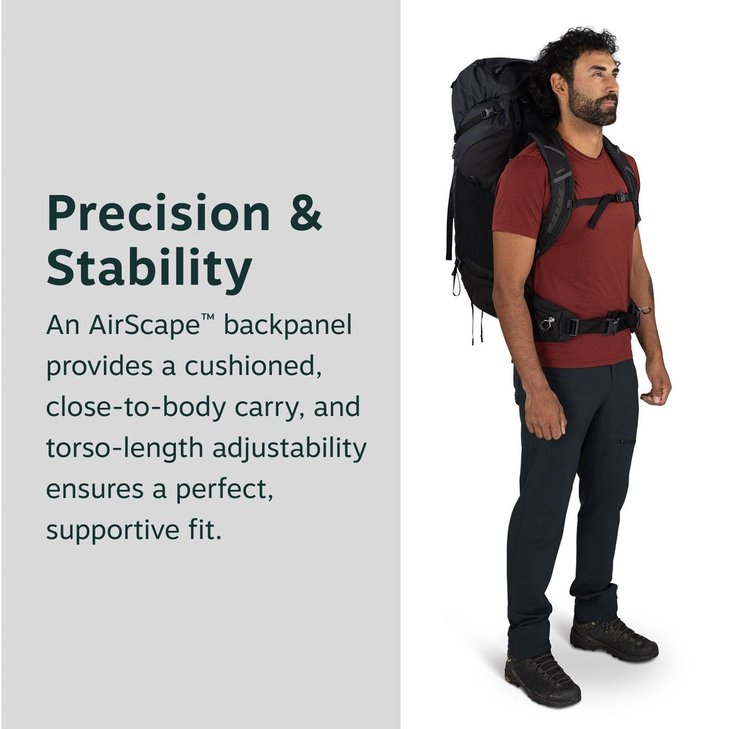 The image shows a man standing and wearing a large, black hiking backpack. He is dressed in outdoor attire, featuring a red short-sleeved t-shirt and dark blue or black pants along with black hiking shoes. The backpack is strapped tightly to his back, sho
