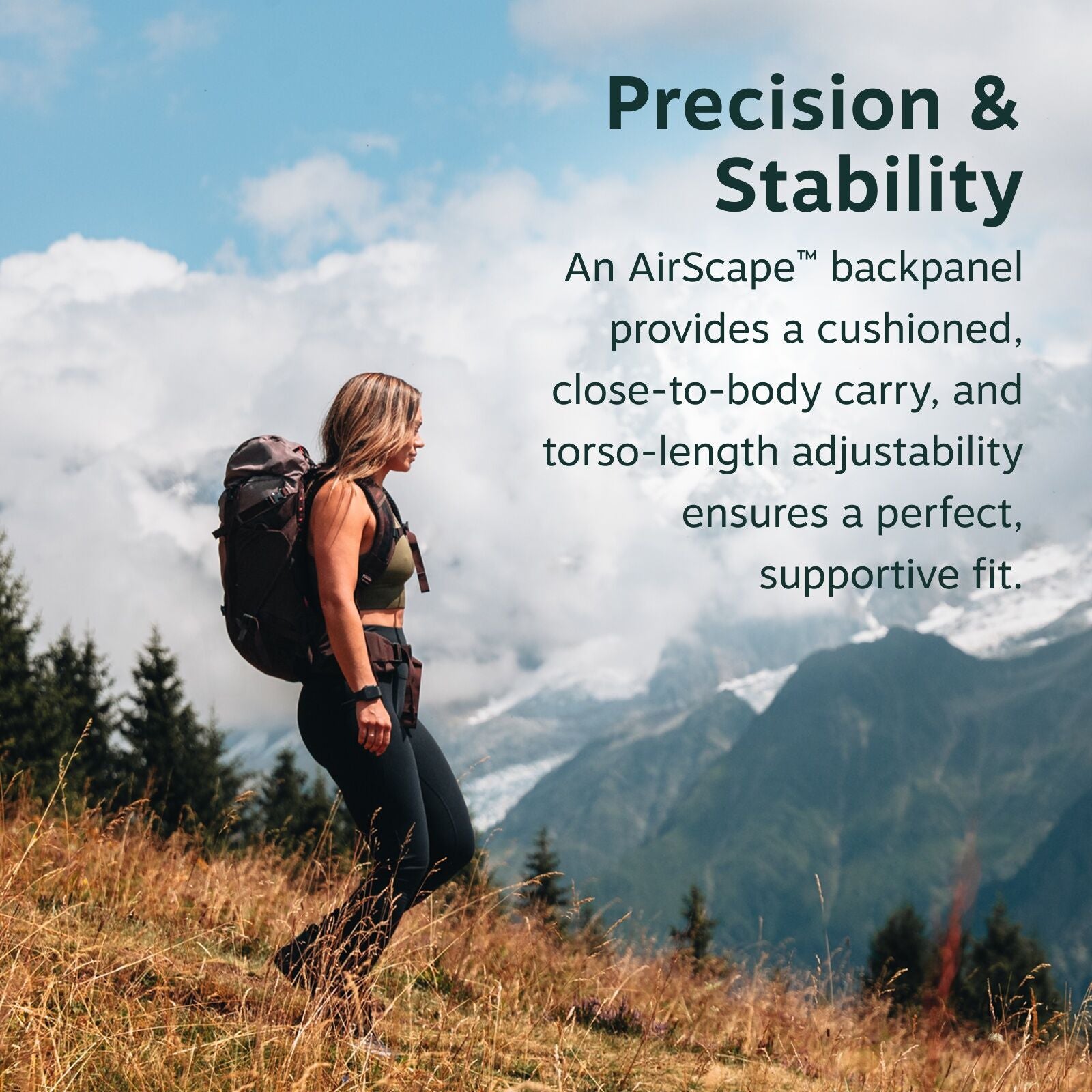 The image features a woman hiking in a scenic mountain landscape. She is wearing a green top and black leggings, carrying a large backpack with a cushioned backpanel designed for a close-to-body carry. The scene captures her walking along a grassy slope,