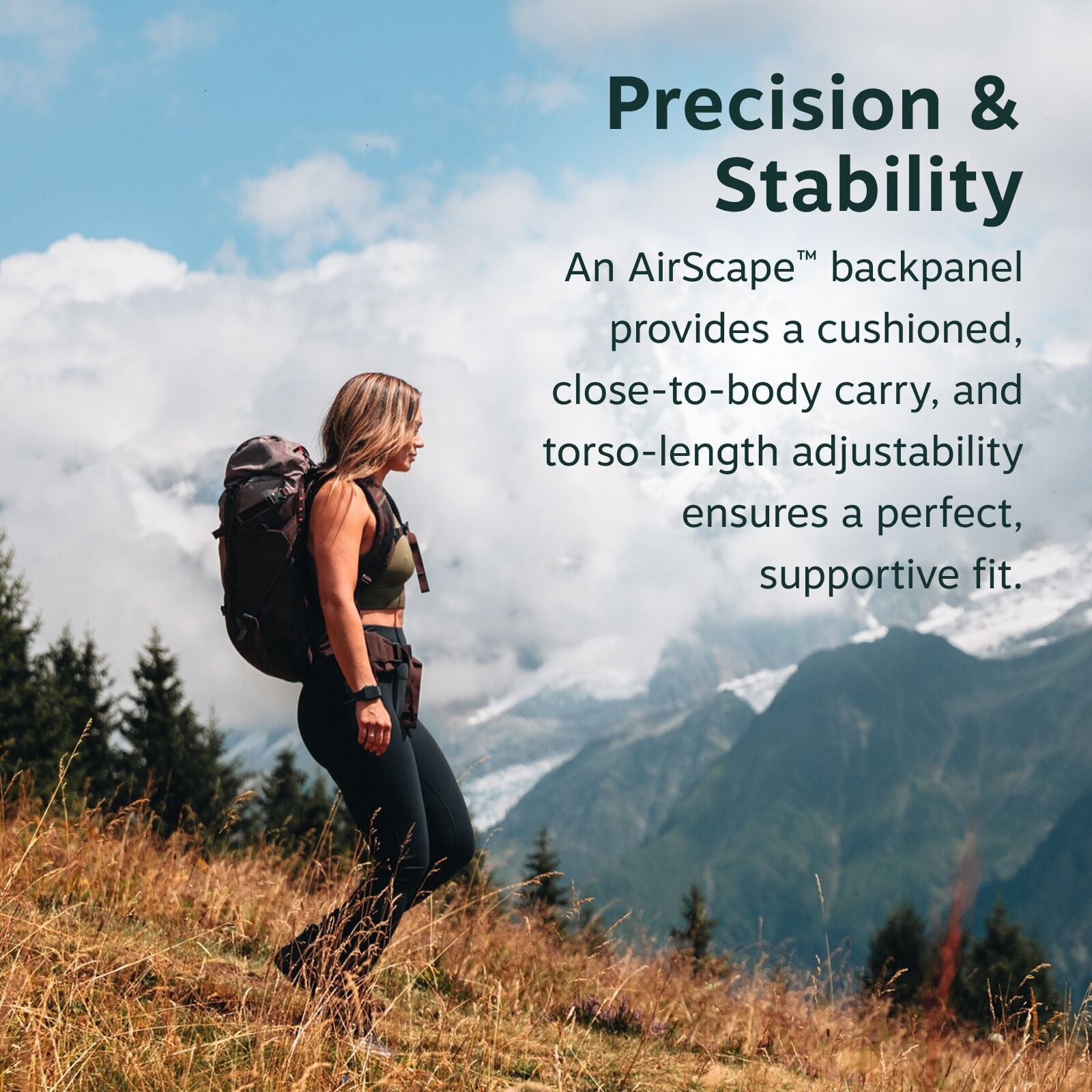 The image features a woman hiking in a scenic mountain landscape. She is wearing a green top and black leggings, carrying a large backpack with a cushioned backpanel designed for a close-to-body carry. The scene captures her walking along a grassy slope,