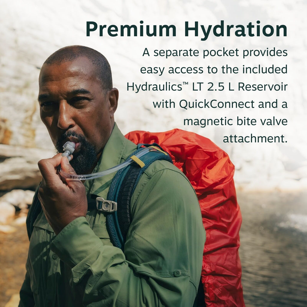 The image features an outdoor setting, with a person holding a drinking tube of a hydration system. The individual is wearing a green long-sleeve shirt, possibly suited for hiking or outdoor activities, along with a backpack. The backpack is equipped with