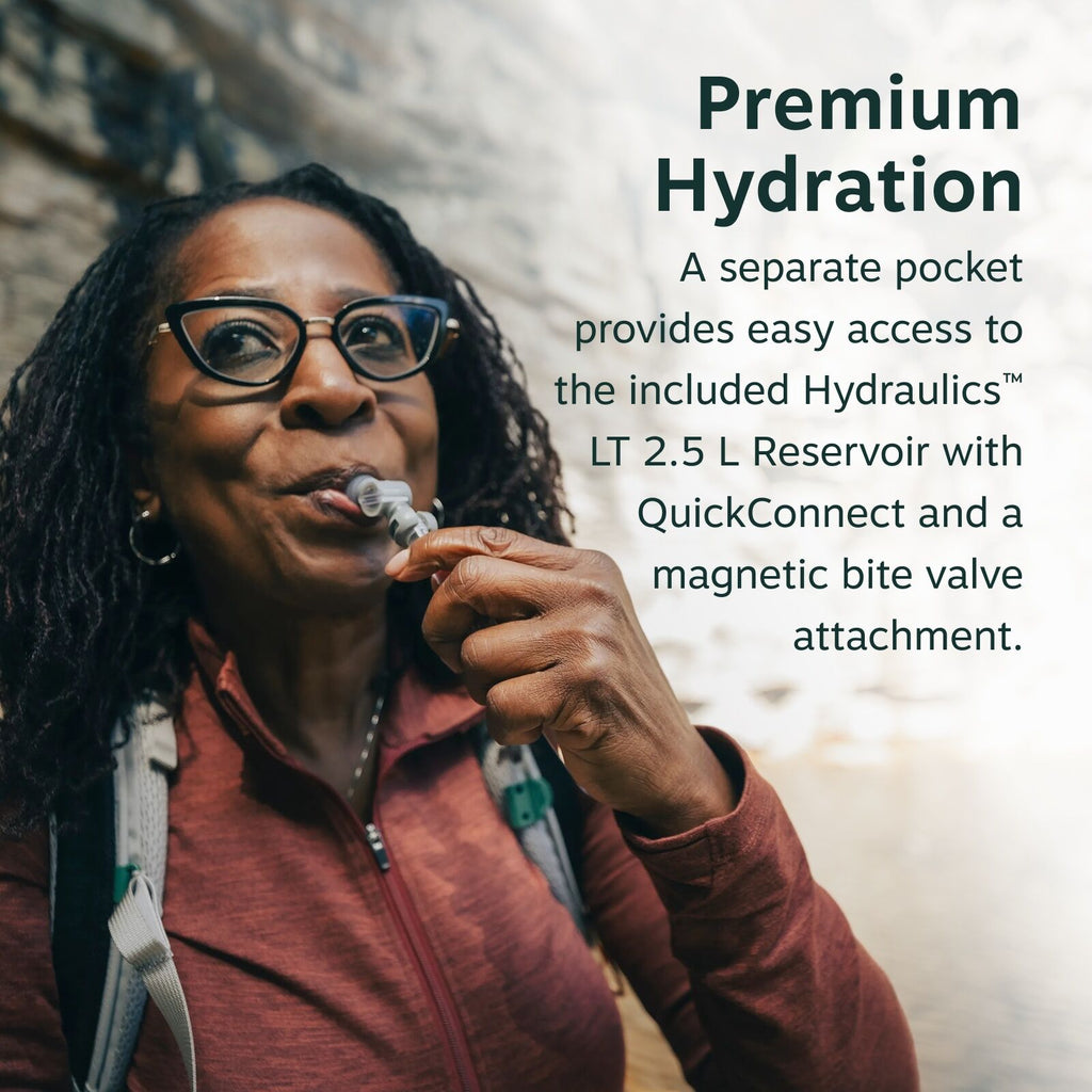 The image depicts a person wearing outdoor clothing, holding a hydration tube, likely from a hydration pack, near their mouth. The individual is wearing a maroon zip-up jacket and a backpack with grey straps, standing in an outdoor setting that resembles