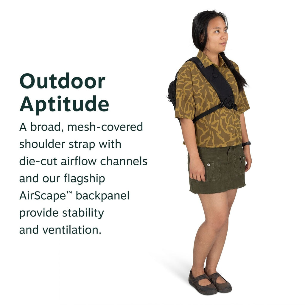 The image features a person standing and wearing a backpack with a broad, mesh-covered shoulder strap. The person's outfit includes a patterned shirt in shades of brown with an abstract design, a dark green mini skirt with pockets, and a pair of black sho