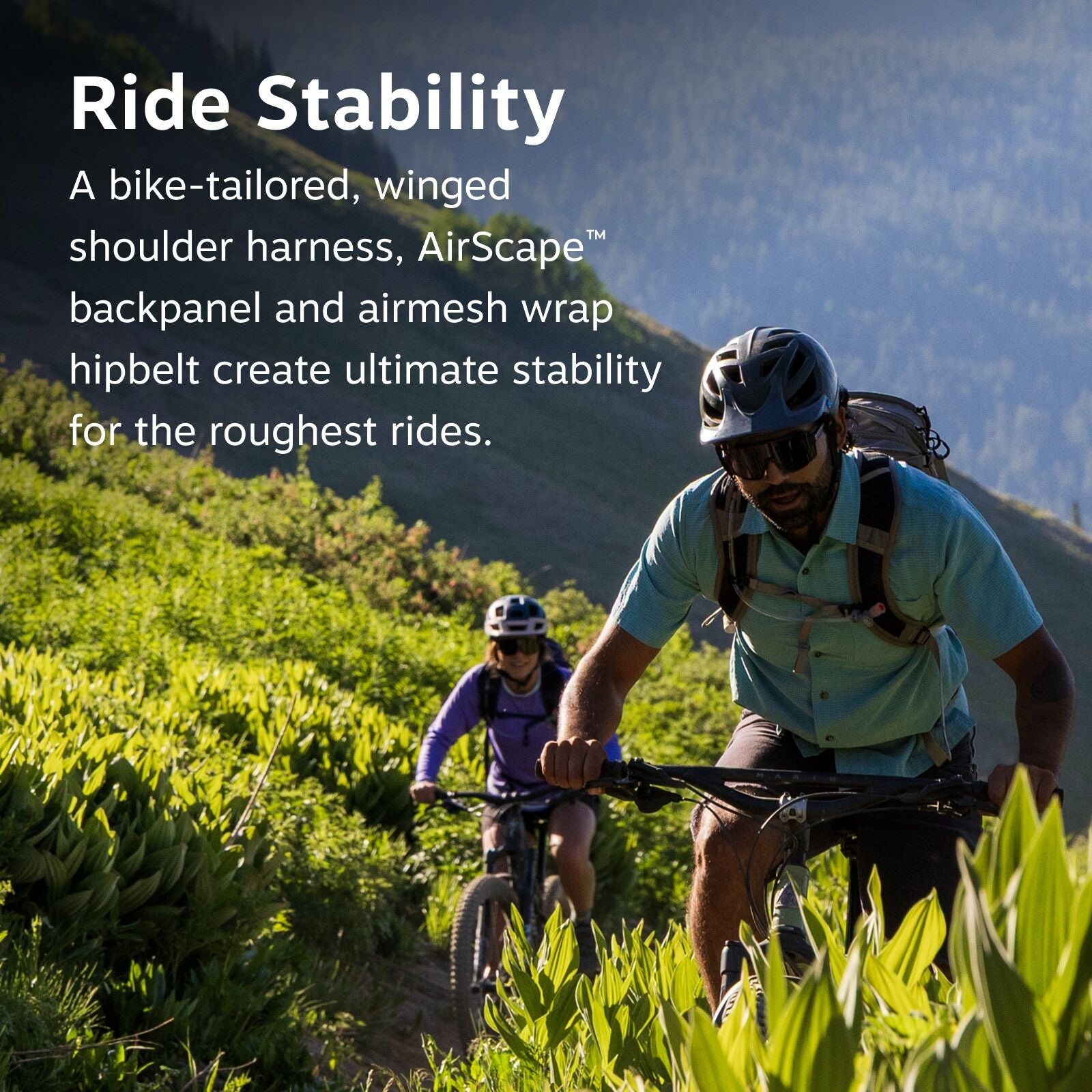 The image features two individuals mountain biking on a lush, green trail surrounded by vibrant foliage. The person in the foreground is wearing a light blue shirt and a helmet while riding their bicycle confidently. Both cyclists are equipped with backpa