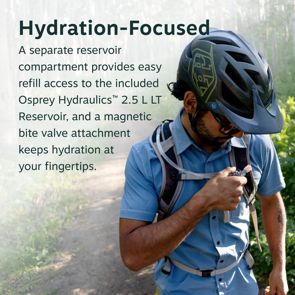 The image depicts a person wearing outdoor gear on a trail surrounded by lush greenery. The individual is dressed in a short-sleeved blue button-up shirt and is sporting a black cycling helmet with a distinctive logo on the side. They are equipped with a