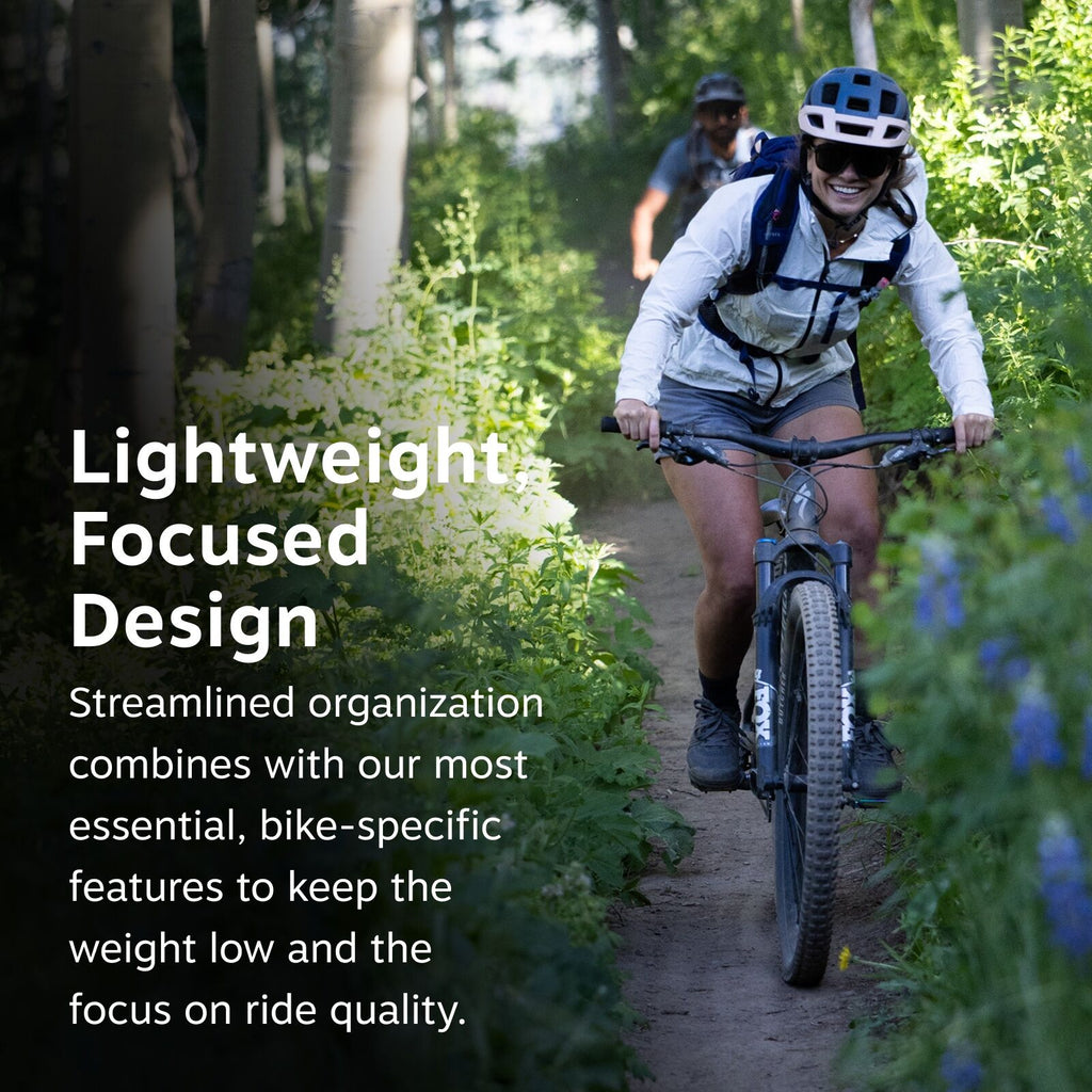 The image features a person riding a mountain bike along a narrow dirt trail that winds through a lush, green forest. They are dressed in a white jacket, dark shorts, and a helmet, suggesting they are equipped for a biking adventure. The path is surrounde