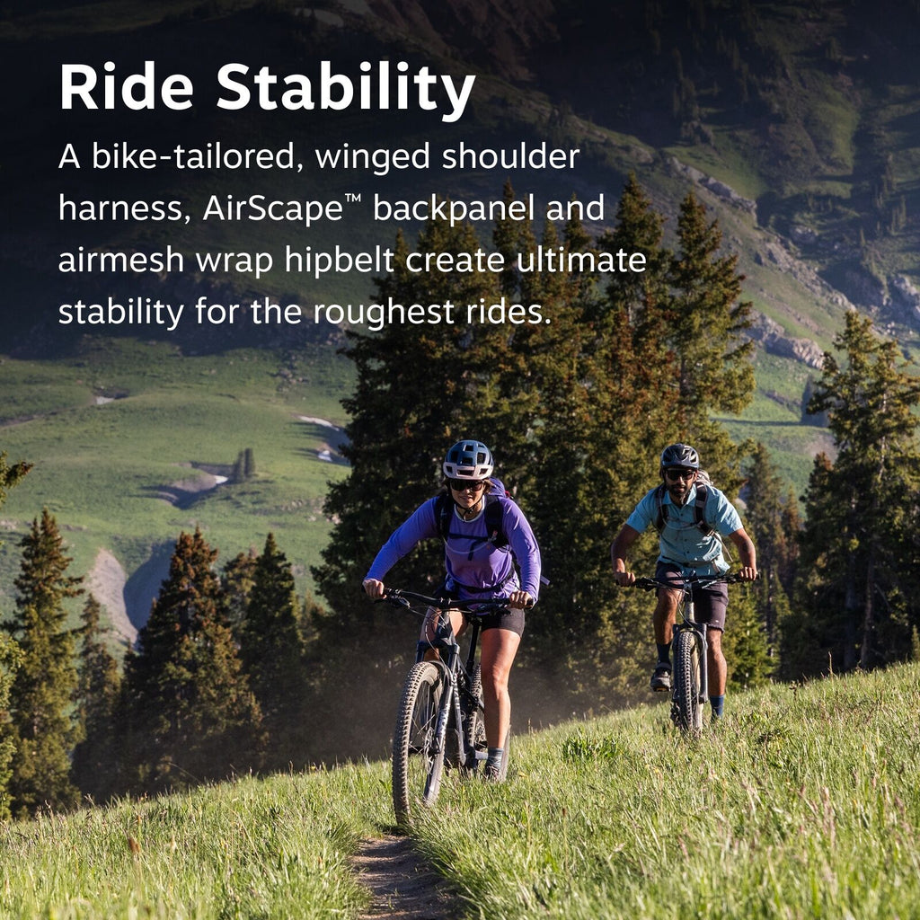 The image captures two cyclists riding mountain bikes on a grassy trail in a picturesque mountainous area. The trail is narrow and flanked by trees and grass, with a backdrop of lush green hills and cliffs. The cyclist in the foreground is wearing a purpl