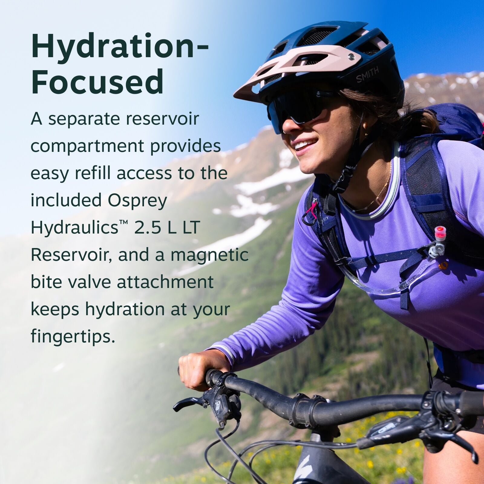 The image features a person riding a mountain bike, focusing on the use of a hydration pack. They are wearing a purple long-sleeve shirt and a helmet, with a hydration tube accessible on their chest. The background shows a mountainous landscape with blue