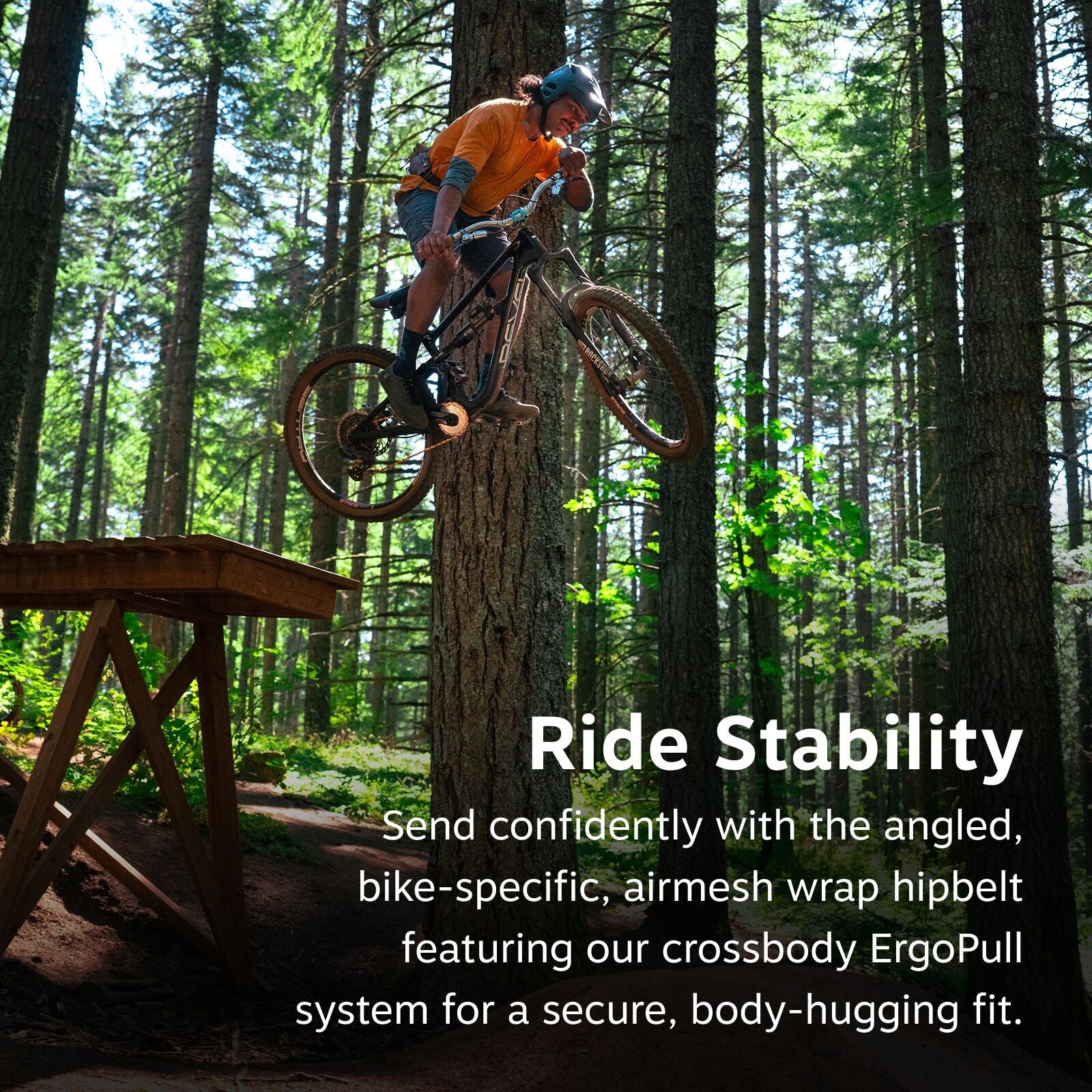 The image captures an exhilarating moment of a mountain biker in mid-air after launching off a wooden ramp in a forested area. The rider is wearing a blue helmet, an orange long-sleeve shirt, and shorts, skillfully maneuvering a bike labeled "POC". The ba