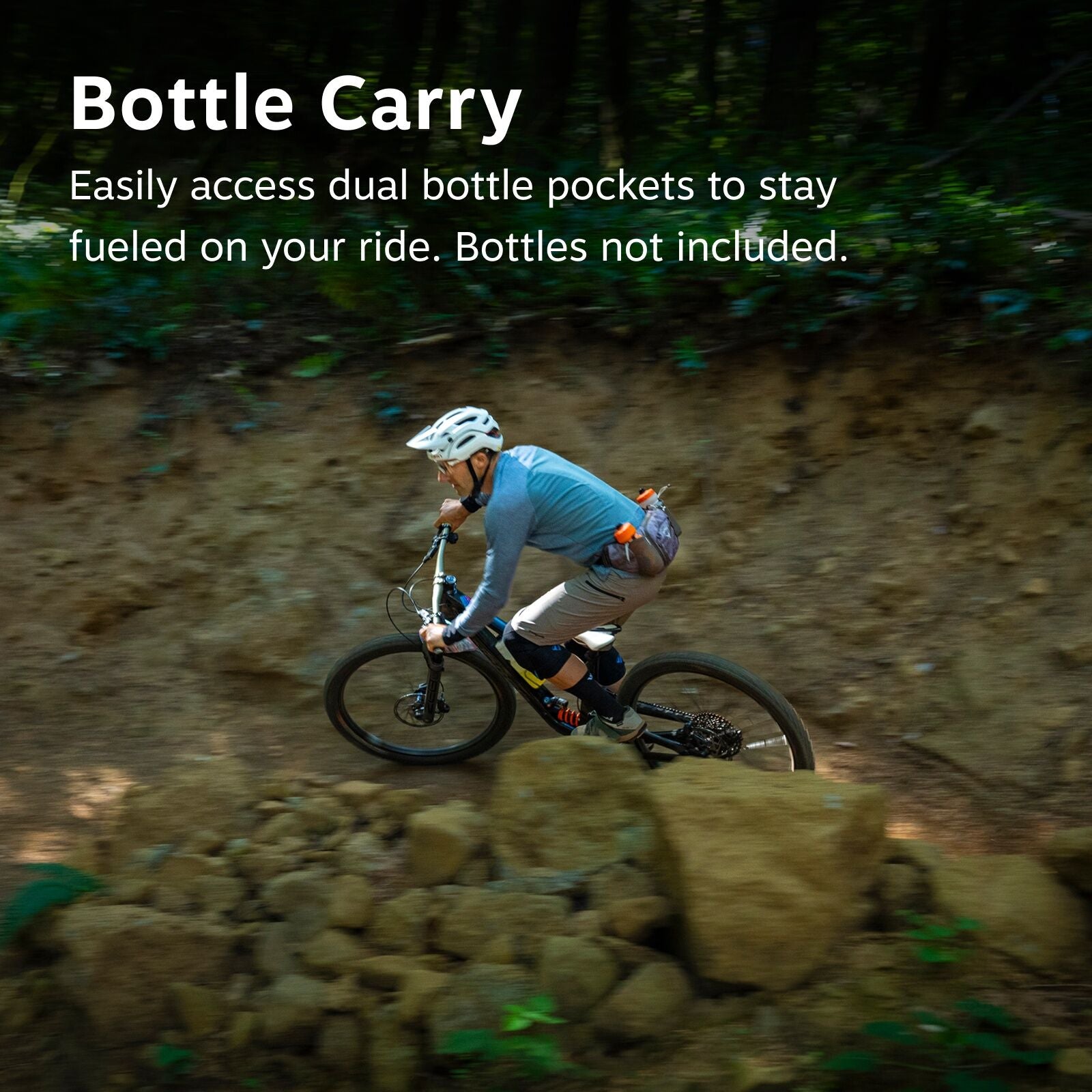 The image depicts a mountain biker navigating a rugged trail, surrounded by a lush, green forest backdrop. The biker is wearing a white helmet and a long-sleeve blue shirt, paired with beige shorts and knee pads for safety. A small grey pack is strapped t