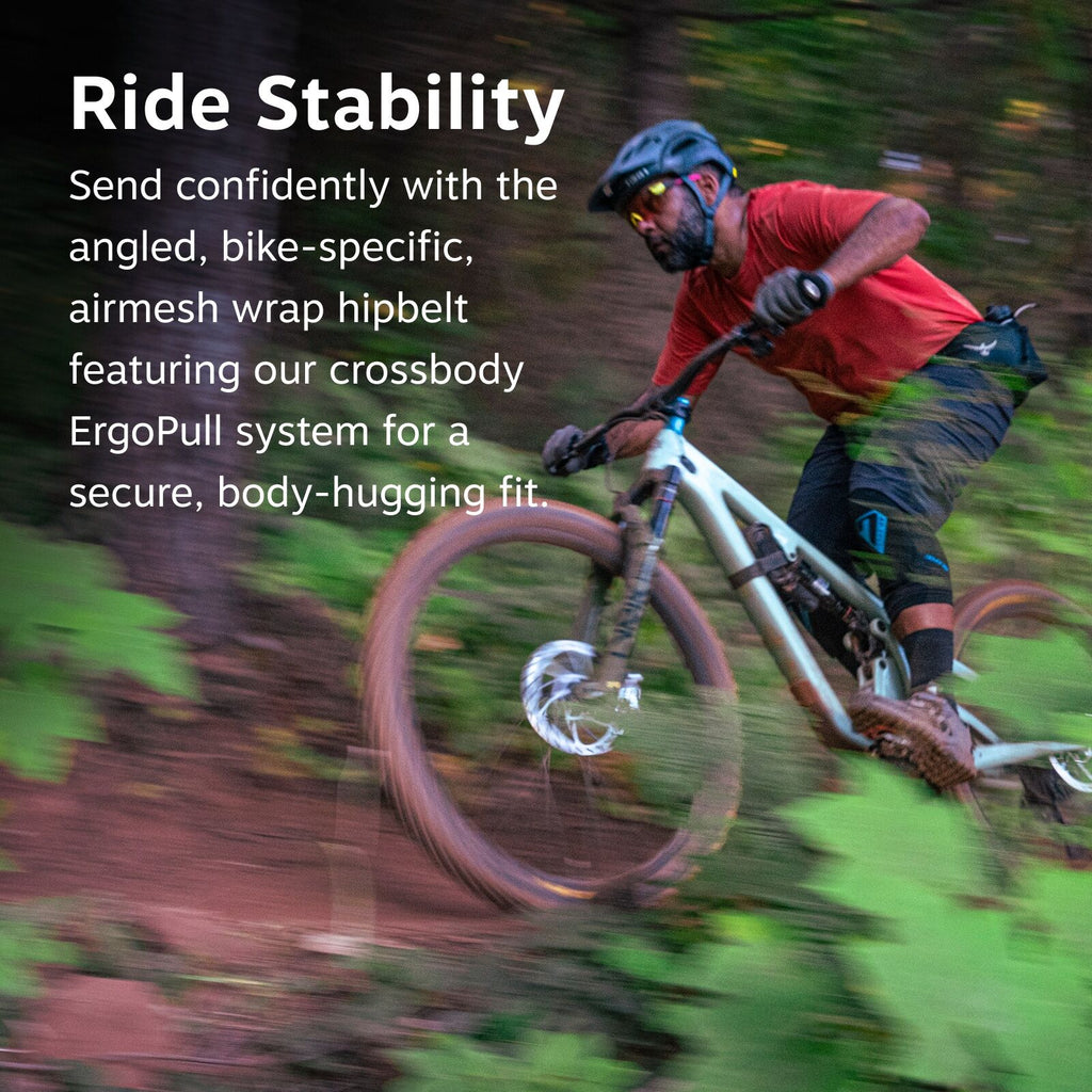 The image features an action shot of a mountain biker riding downhill through a forest. The biker is wearing a bright red shirt, black pants, and protective gear, including knee pads and a helmet. The bike is white, and the rider appears to be in motion,