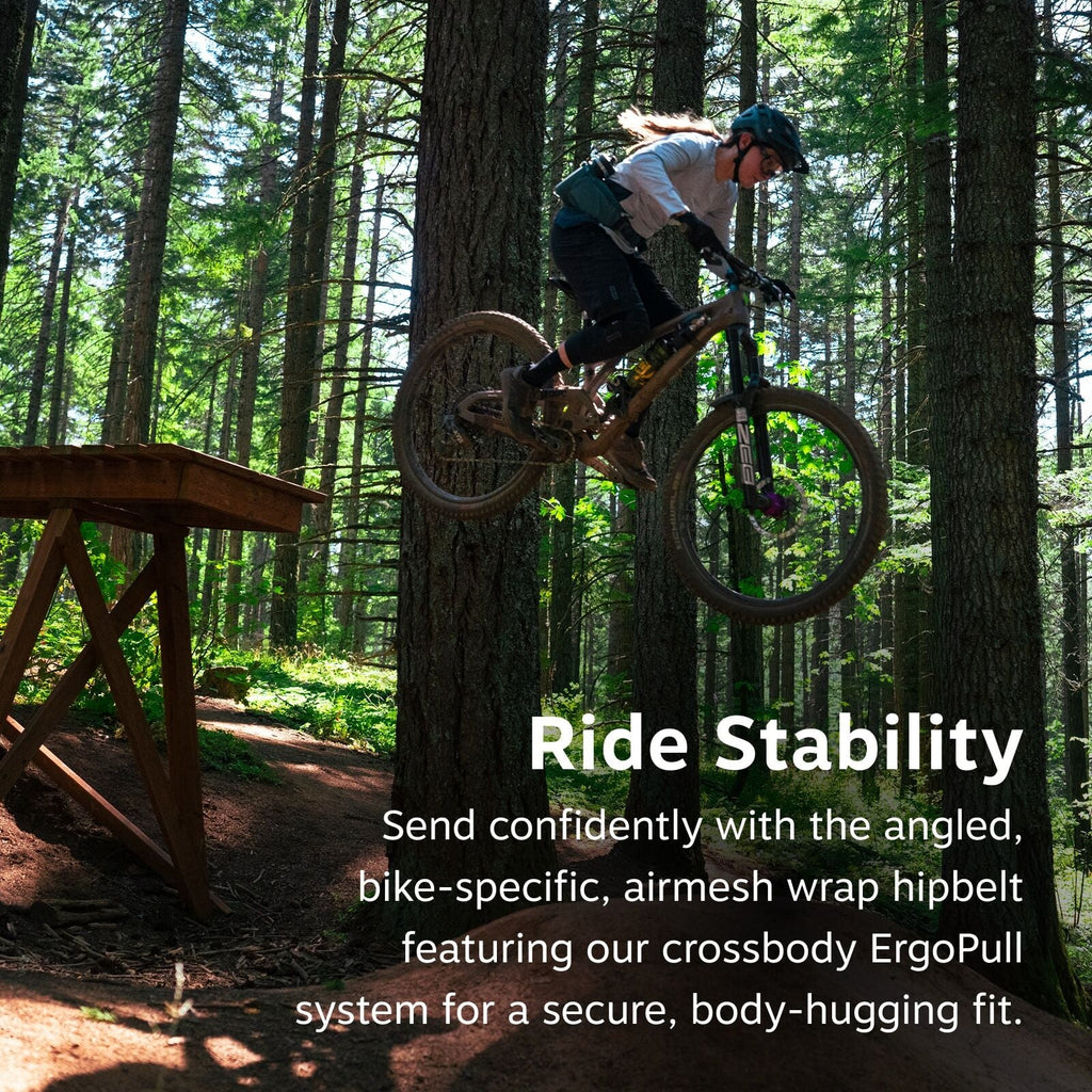 The image captures a dynamic moment of a mountain biker airborne, having just launched off a wooden ramp in a forest setting. The rider is wearing protective gear, including a helmet and gloves, and is equipped with a teal-colored backpack or hip belt. Th