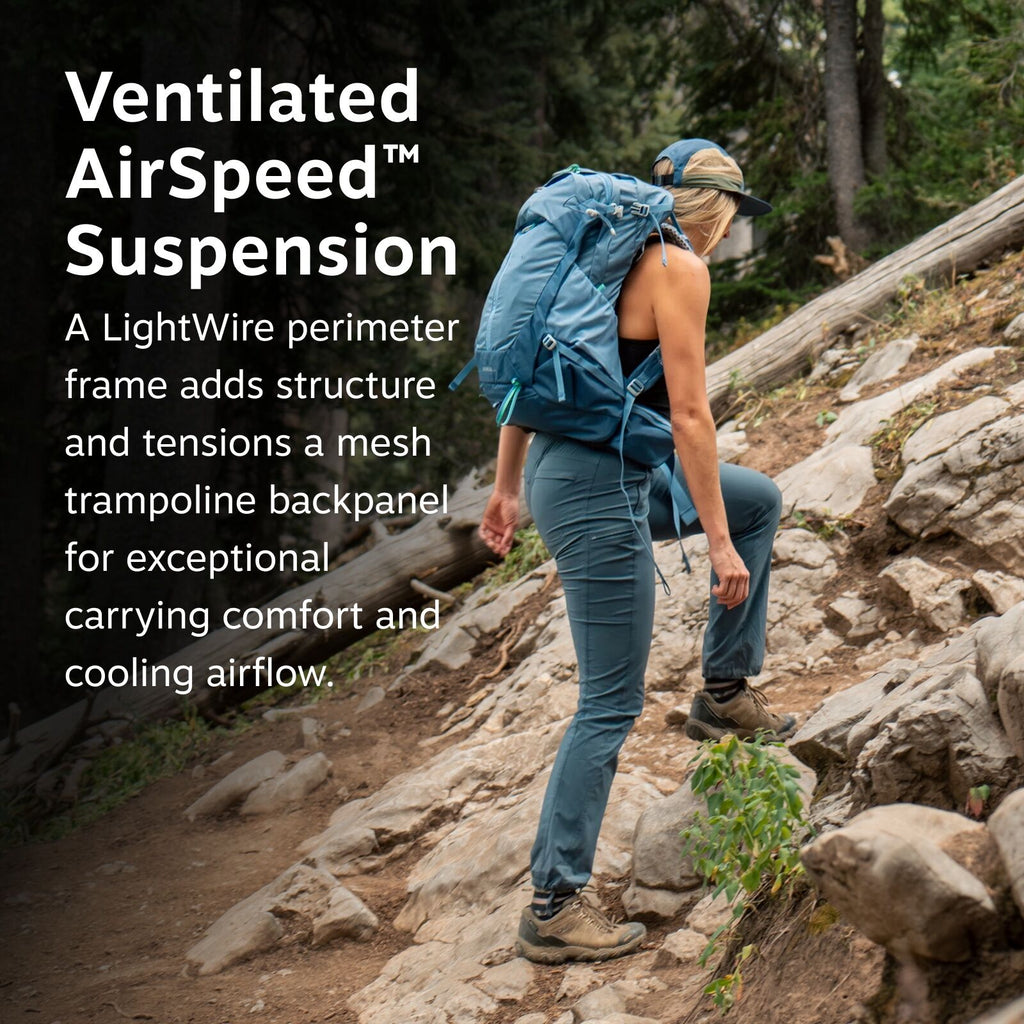 The image shows a woman hiking up a rocky trail in a forested area, wearing a blue backpack. She is dressed in blue outdoor pants, a black sleeveless top, hiking boots, and a dark cap. The trail is surrounded by trees and rocks, indicating a rugged outdoo