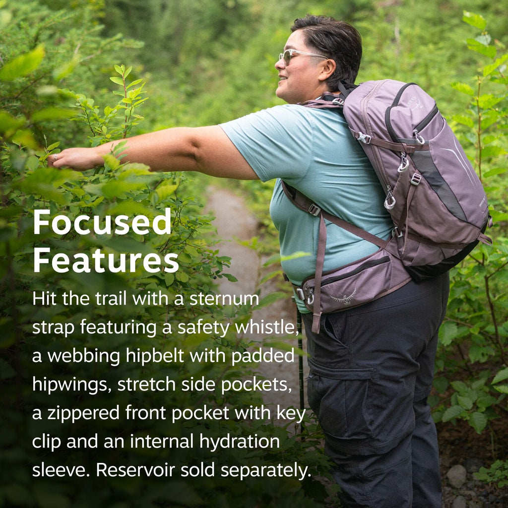 The image depicts a person standing on a hiking trail surrounded by lush greenery. The individual is wearing a light blue t-shirt and dark pants while carrying a large gray and purple hiking backpack. The backpack is equipped with various features such as