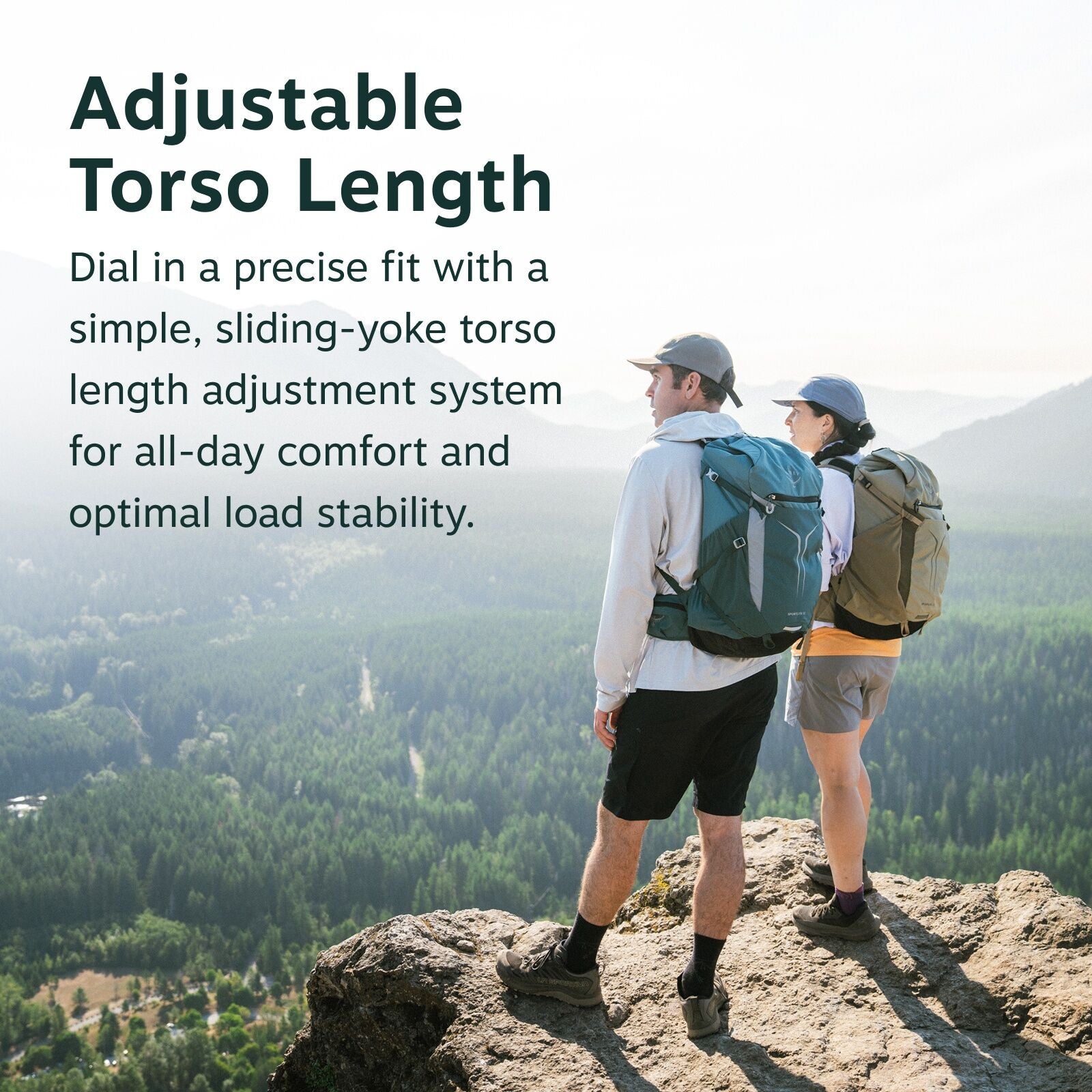 The image features two hikers standing on a rocky cliff overlooking a scenic landscape filled with lush green forests and distant mountain ranges. Both individuals are wearing outdoor attire, including hats and backpacks, as they take in the expansive vie