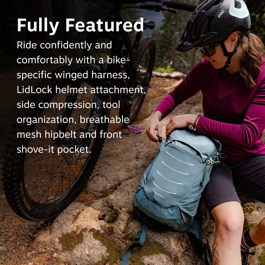 The image depicts a person seated on a rocky terrain near a mountain bike, adjusting a blue backpack labeled "SYNCRO 12" from the brand Osprey. The individual is dressed in a maroon long-sleeve top, black cycling shorts, and is wearing a black cycling hel