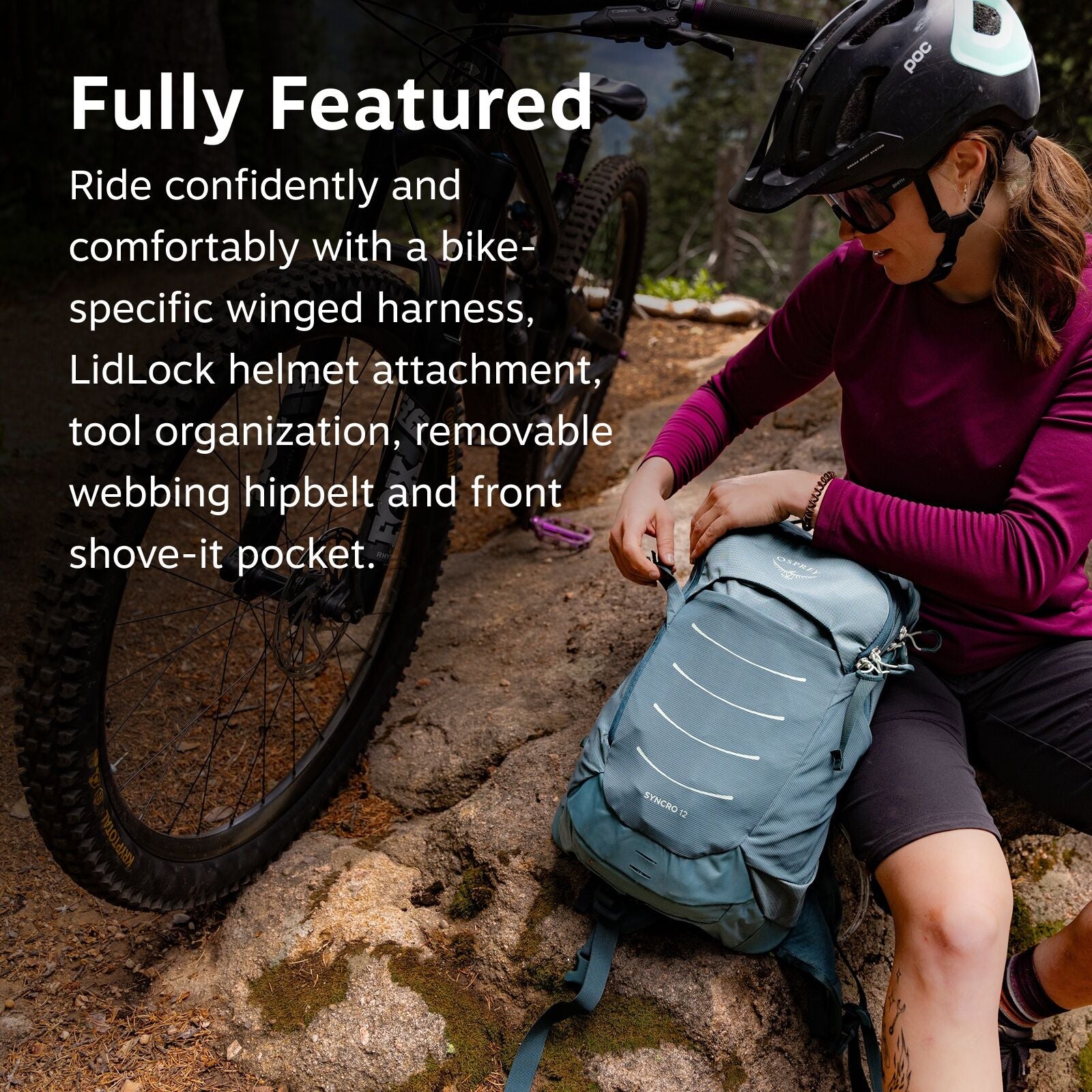 The image shows a woman sitting on a rock next to a mountain bike in a wooded area, likely taking a break from cycling. She is wearing a dark-colored helmet and a long-sleeved, maroon top with black shorts. Her backpack, labeled "SYNCRO 15" by Osprey, is