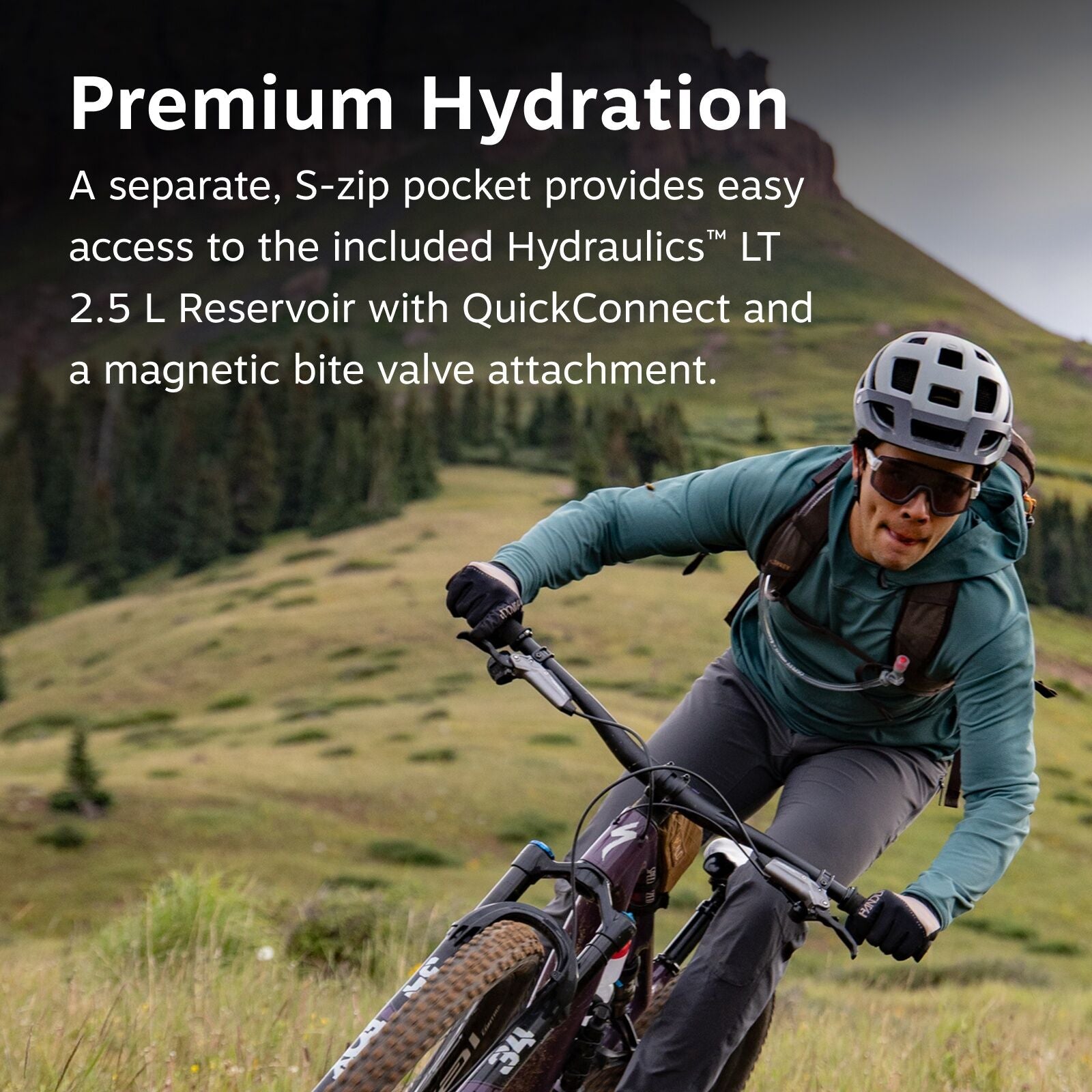 The image features a mountain biker navigating a grassy terrain with a steep, rocky backdrop. The rider is equipped with protective gear, including a white helmet, gloves, and a teal jacket. They are carrying a backpack, which is likely used to hold a hyd