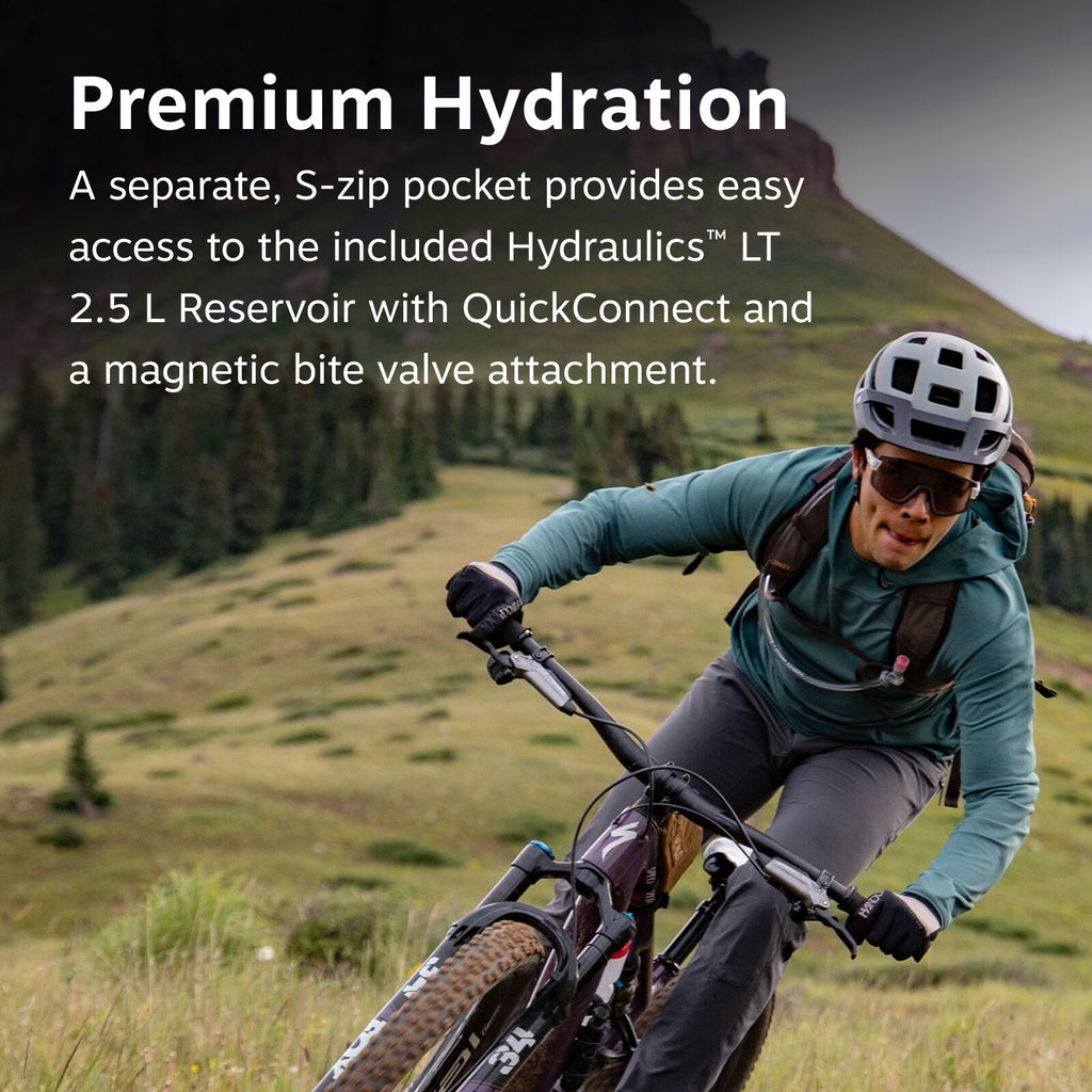 The image features a mountain biker navigating a grassy terrain with a steep, rocky backdrop. The rider is equipped with protective gear, including a white helmet, gloves, and a teal jacket. They are carrying a backpack, which is likely used to hold a hyd