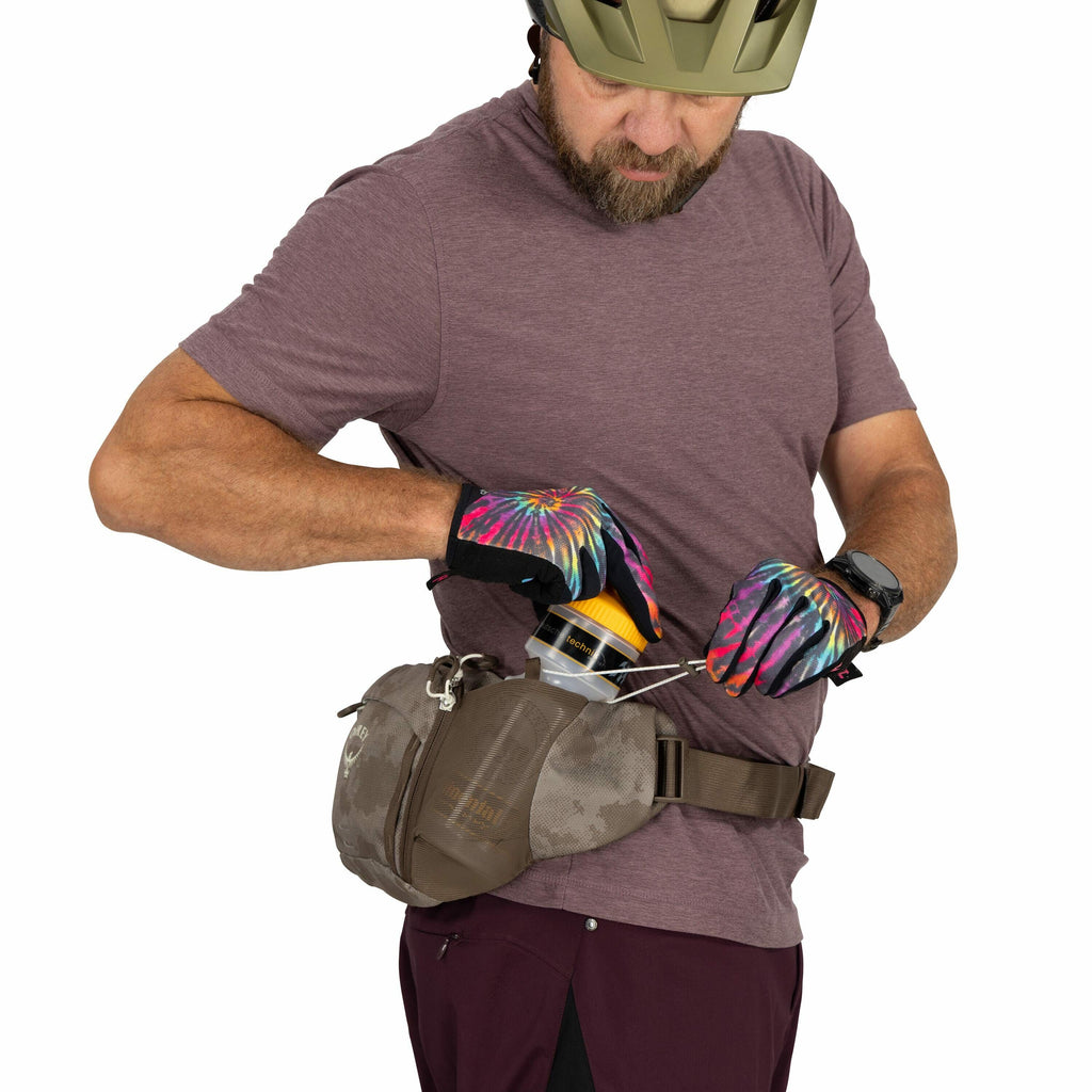 The image depicts a person wearing a brown t-shirt and maroon pants, with a protective helmet on their head. They have a tan waist bag, branded "Osprey," fastened around their waist. The person is using their colorful, tie-dye patterned gloves to handle a