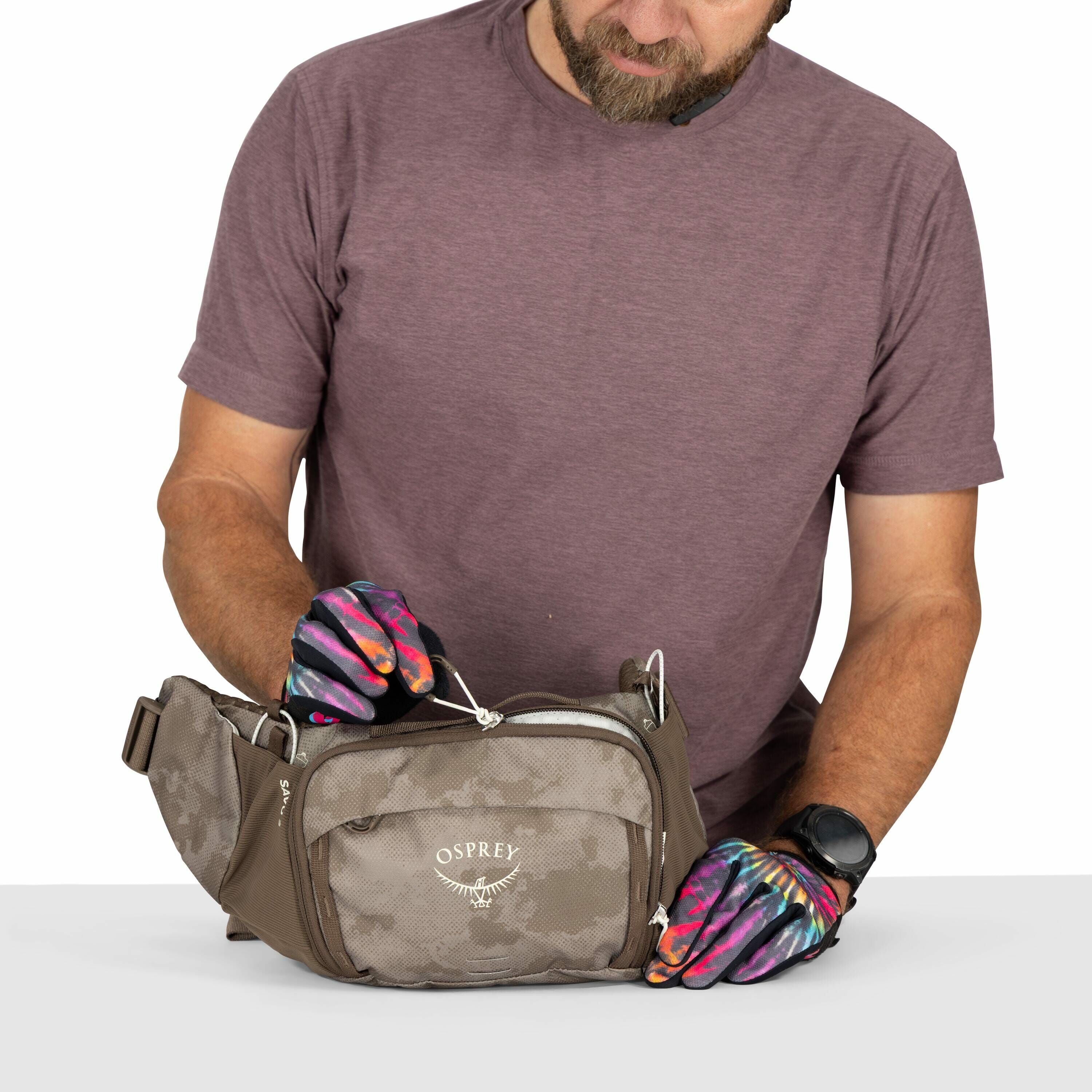 The image features a man in a brownish-purple t-shirt examining a brown and beige Osprey brand waist pack. The waist pack has multiple compartments and zippers, and the man is focusing on one of the zippers, pulling it with his gloved hands. His gloves ar
