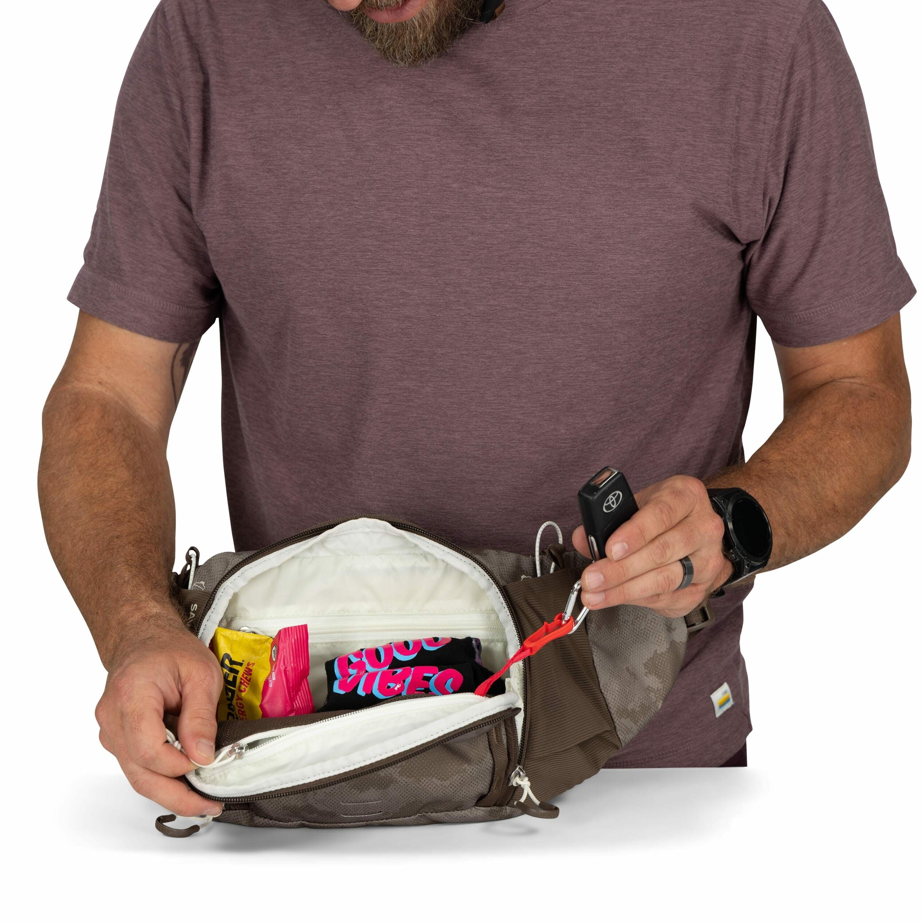 The image shows a man wearing a maroon t-shirt and a black watch, holding an open brown crossbody bag or fanny pack. Inside the bag, there are two packs of Reese's candy, one with a yellow wrapper and the other with a pink wrapper featuring the words "goo