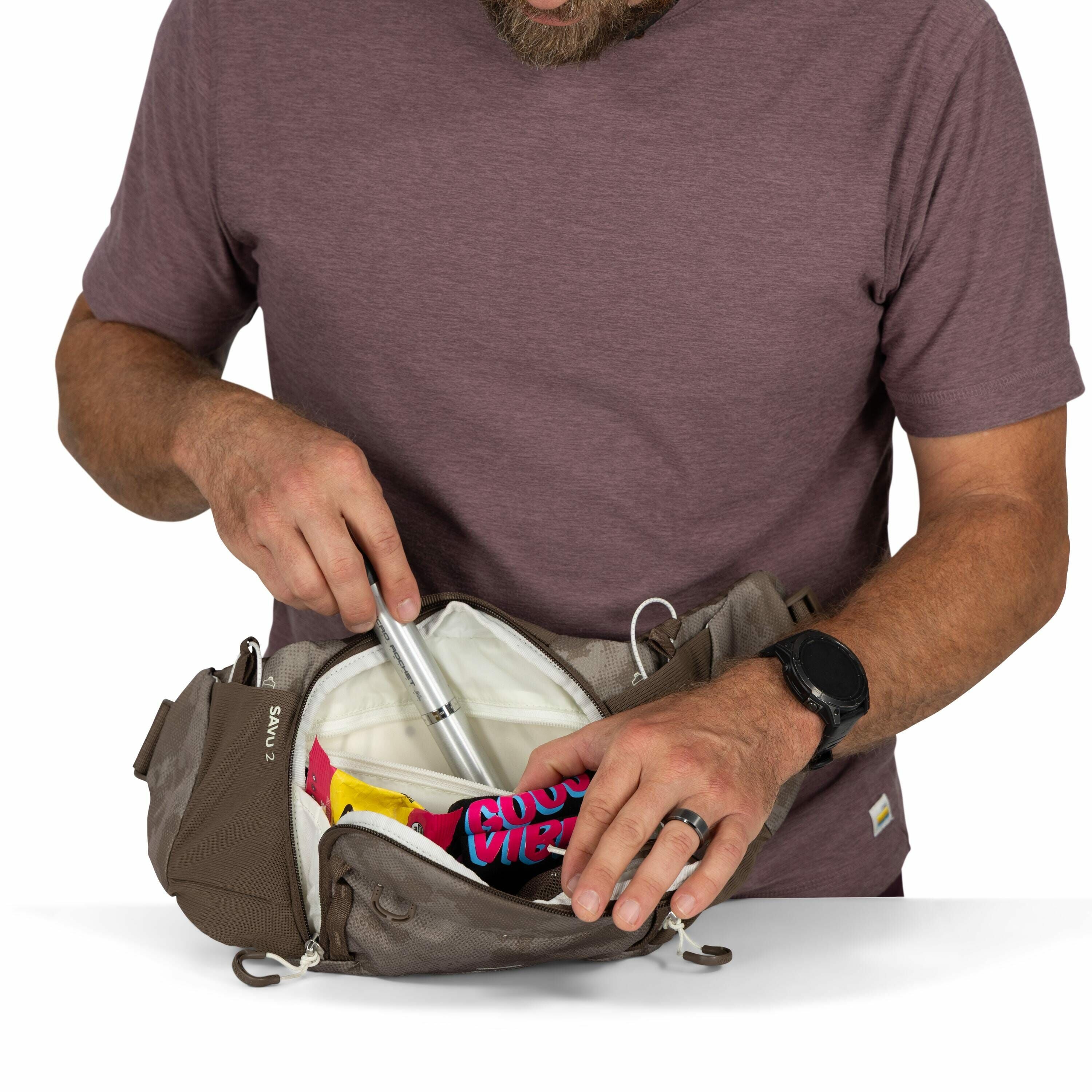The image shows a man wearing a maroon t-shirt who is organizing items in a brown waist pack labeled "Sawtooth 2." He is holding a cylindrical silver object, possibly a pump or tool, and is placing it into the pack. Inside the pack, there is also a colorf